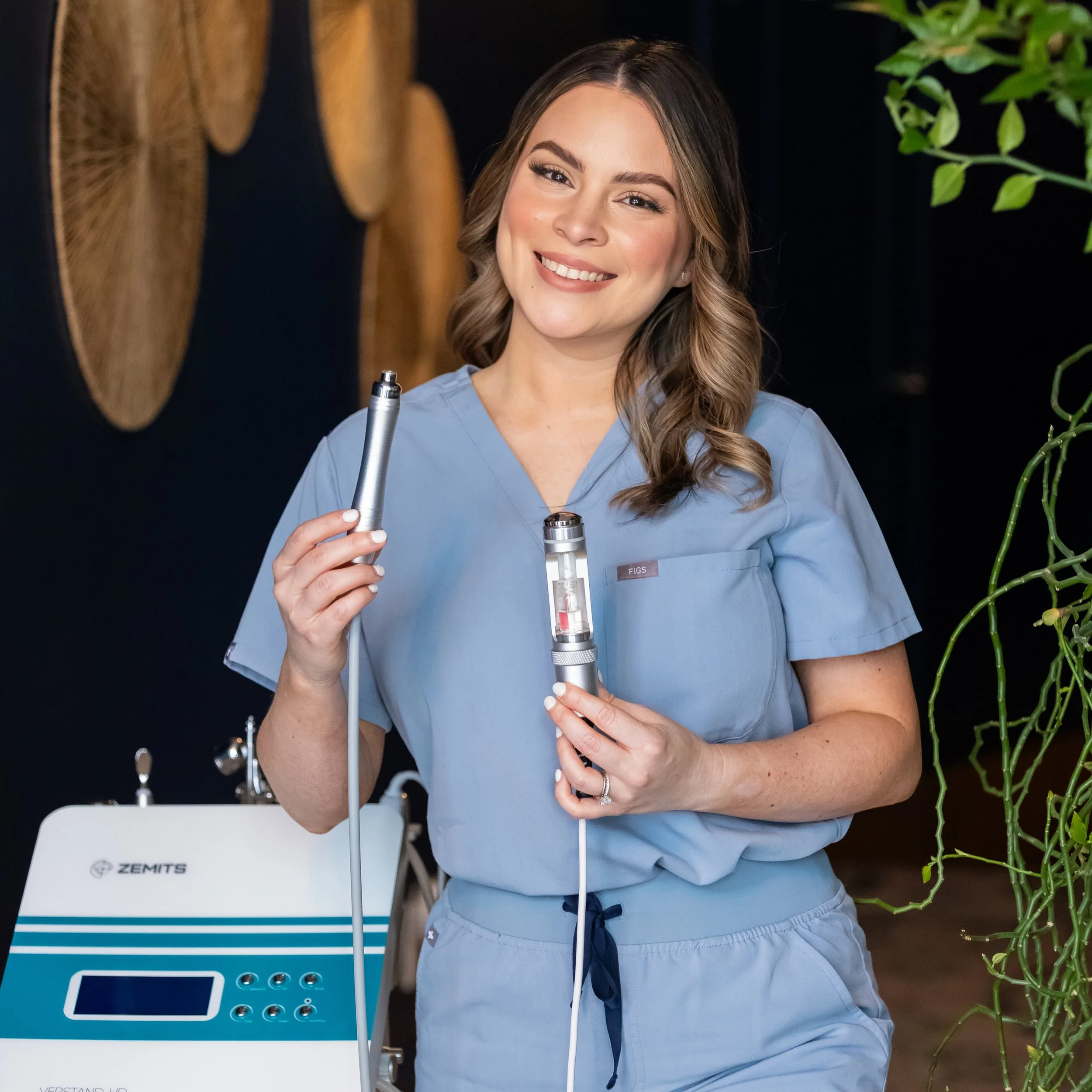 A smiling woman wearing blue medical scrubs holding dental or medical instruments, standing next to a ZEMTS machine in a clinical setting.