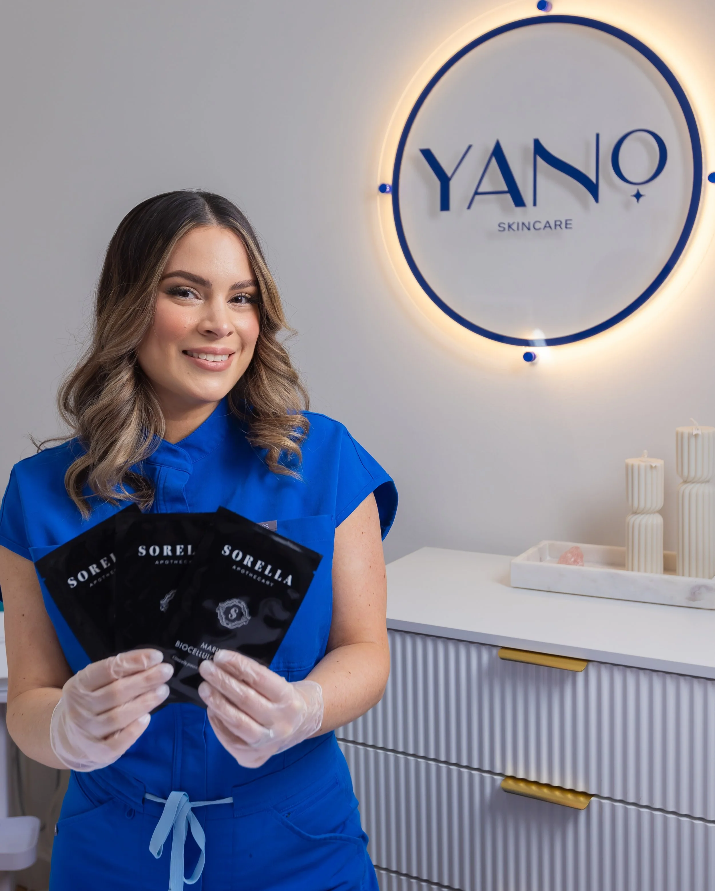 A woman in blue scrubs holding packet samples at YANO Skincare clinic.
