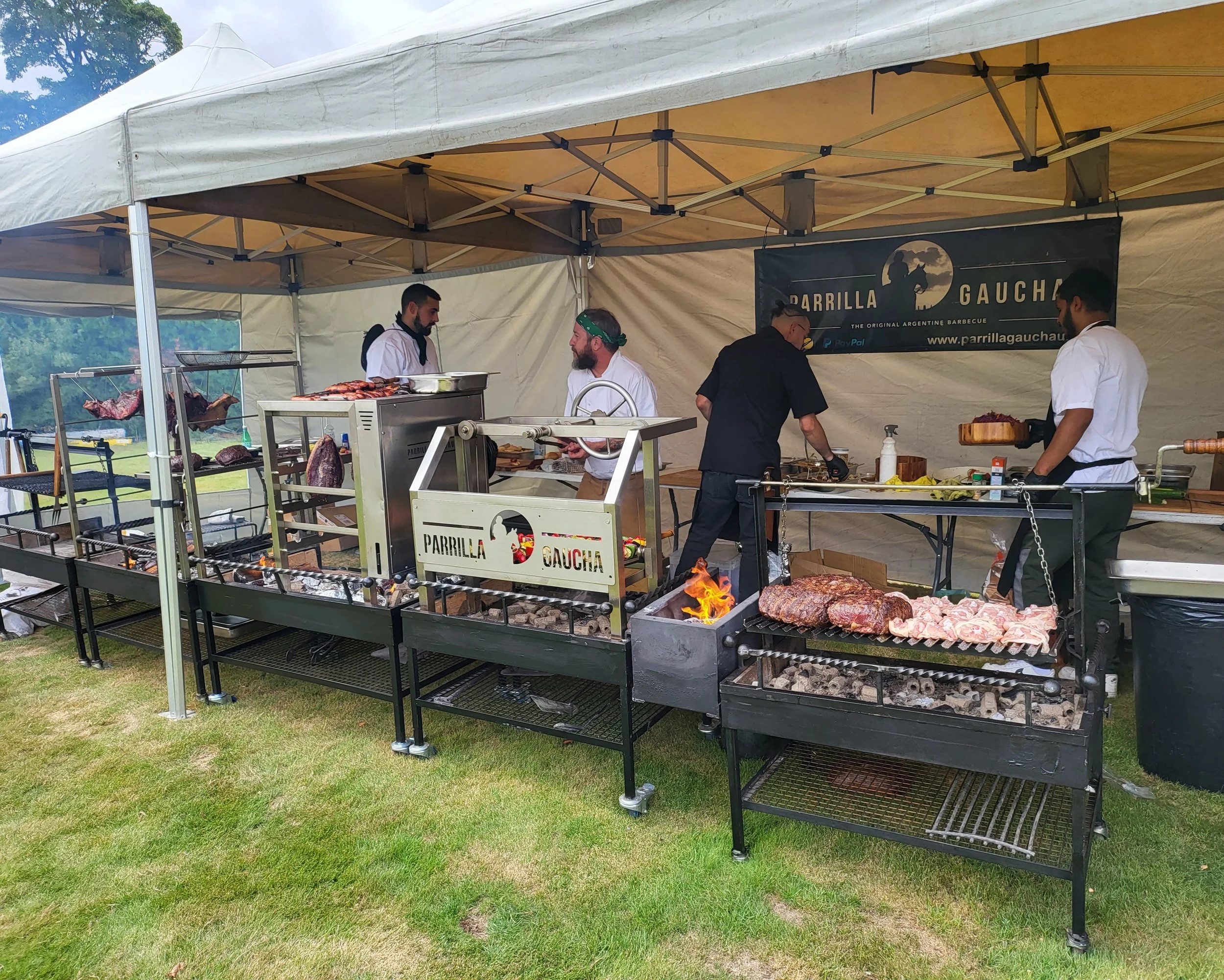 Four men cooking and preparing meat at an outdoor barbecue stand under a canopy, with various grilling equipment and meats on display. A banner reads 'Parrilla Gaucha'.