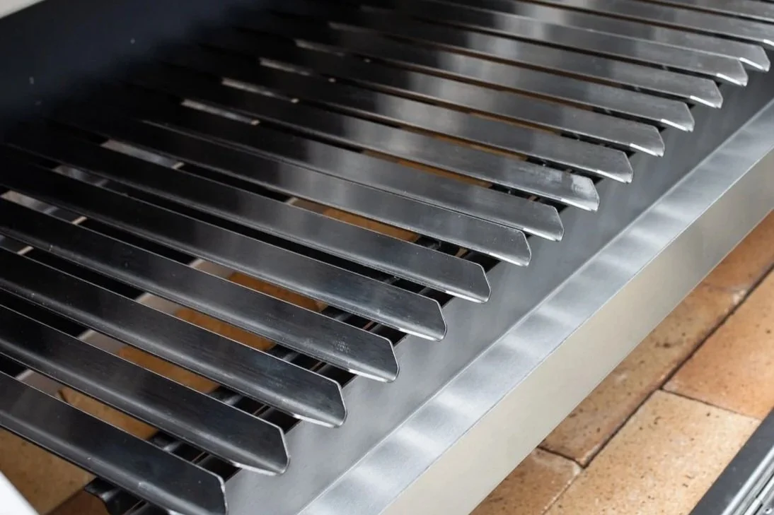 Close-up image of a stainless steel gas grill with black cooking rods and a brick base, viewed from above.