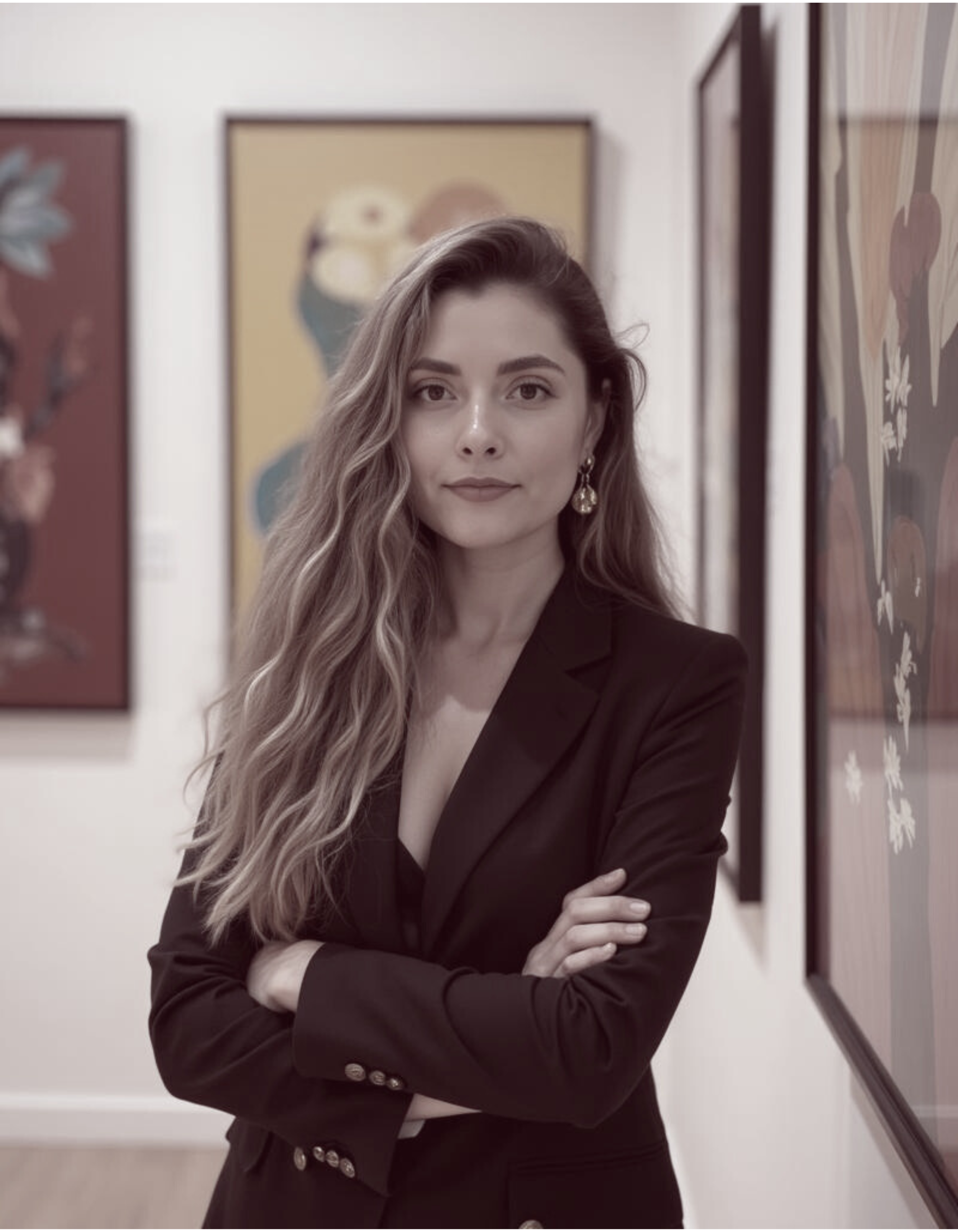 A woman with long wavy hair and earrings standing in an art gallery with her arms crossed, surrounded by colorful artwork.