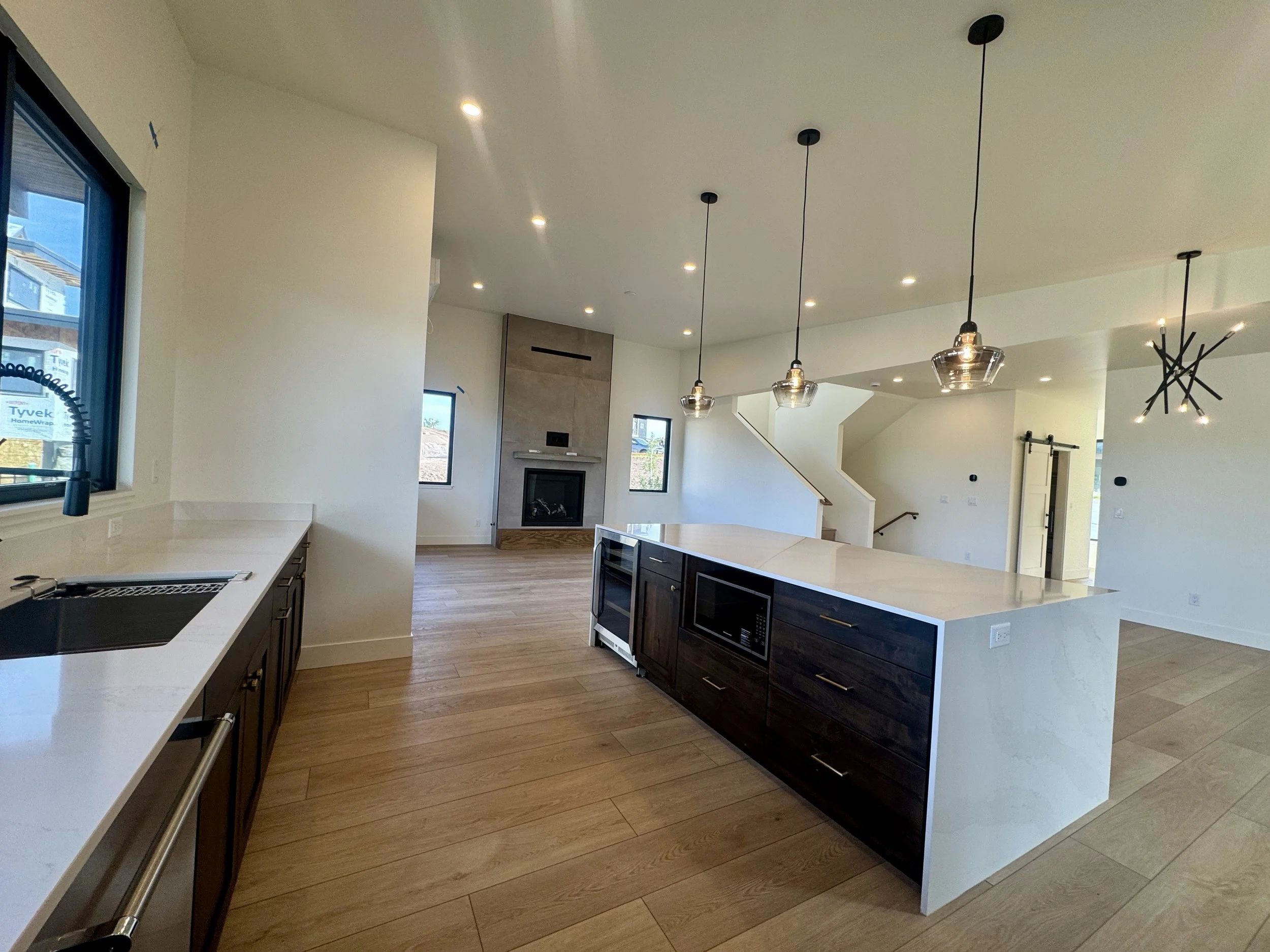 Open-concept modern kitchen with white countertops, dark wood cabinets, stainless steel appliances, pendant lights, and a living area with a fireplace and staircase.