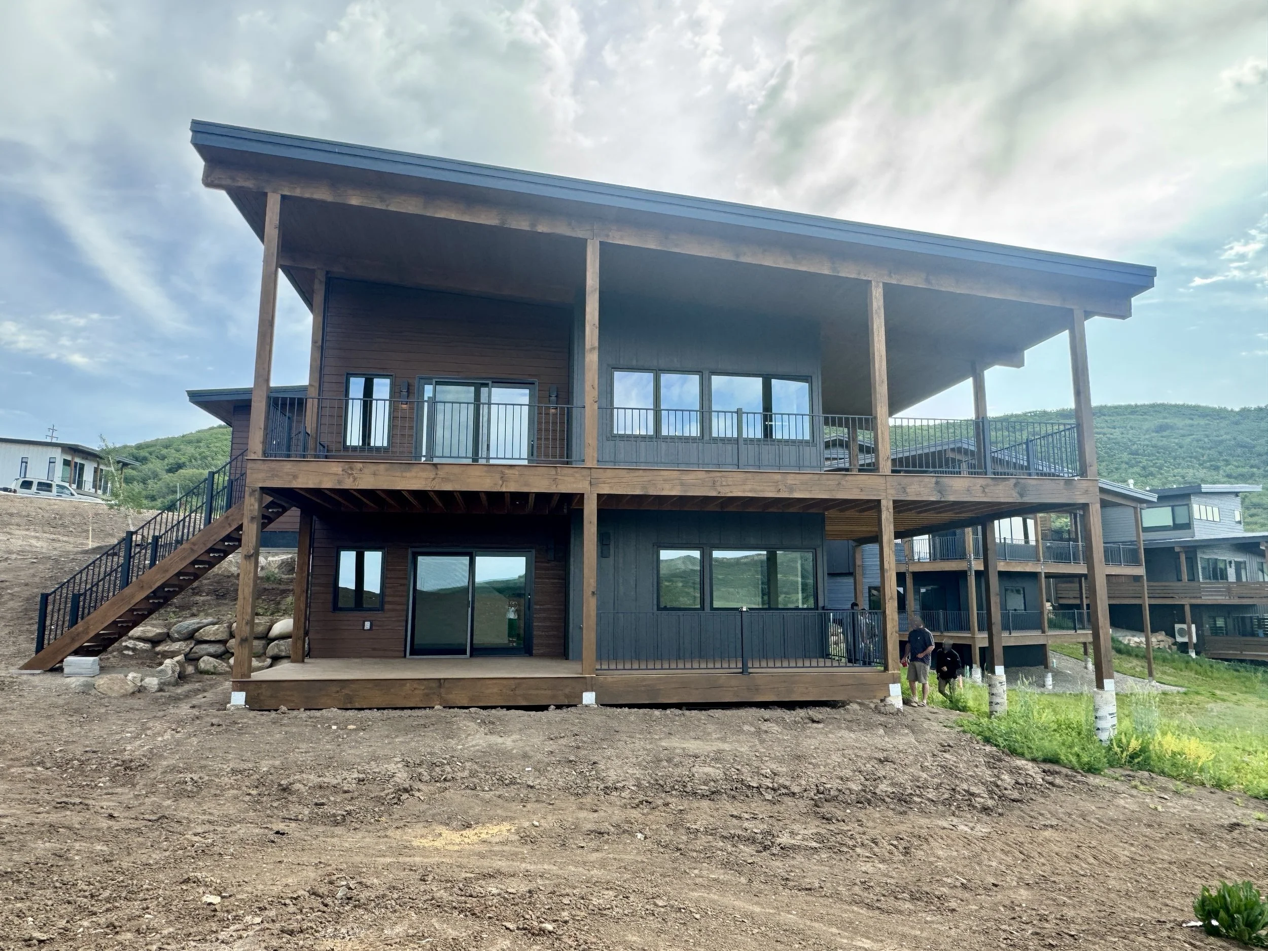 Two-story modern house with a wooden exterior, large windows, and outdoor decking, located on a hillside with a mountain background and partly cloudy sky.