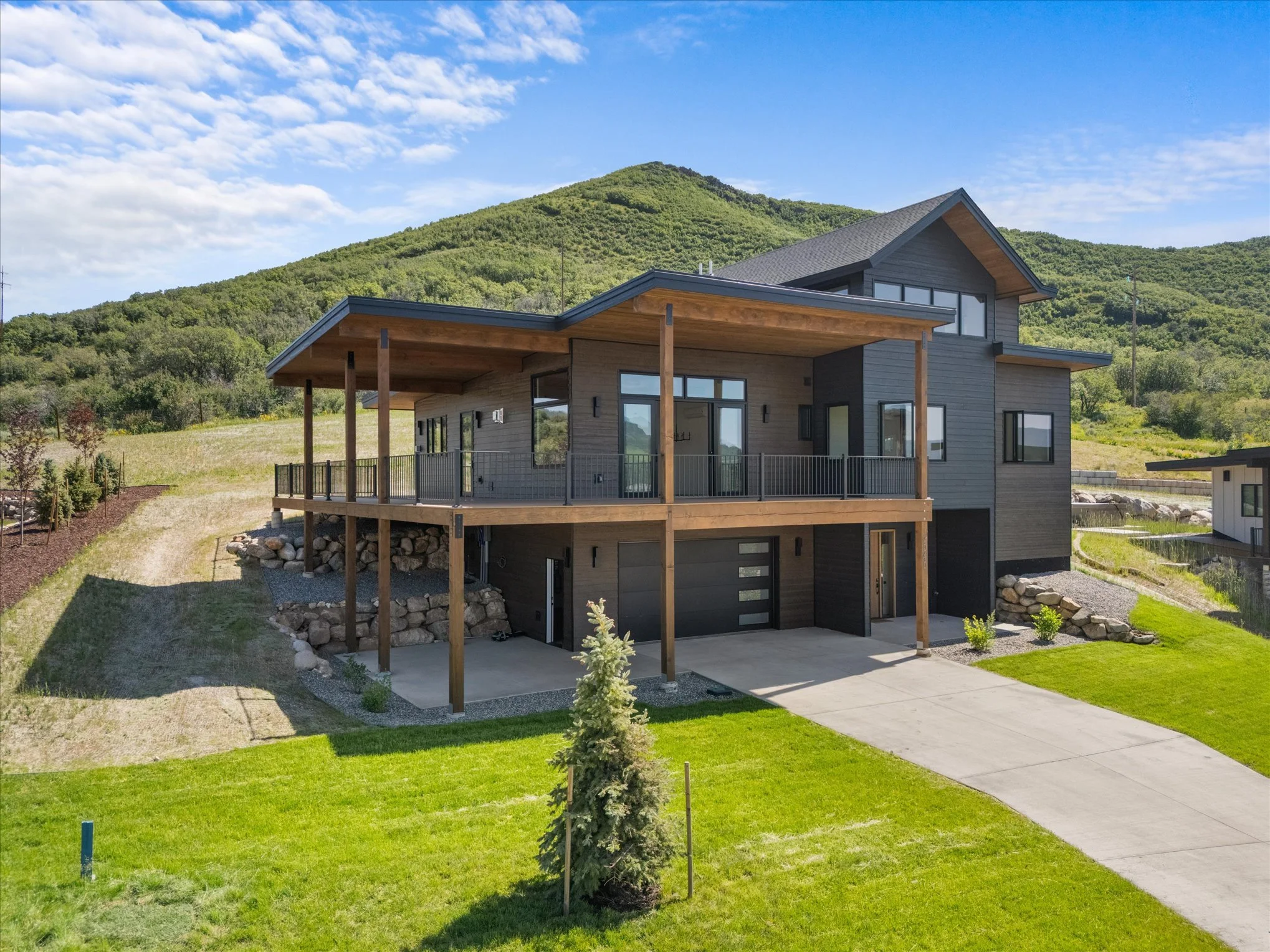 Modern two-story house with a large front deck, gray exterior, and surrounding green lawns and hills under a blue sky.