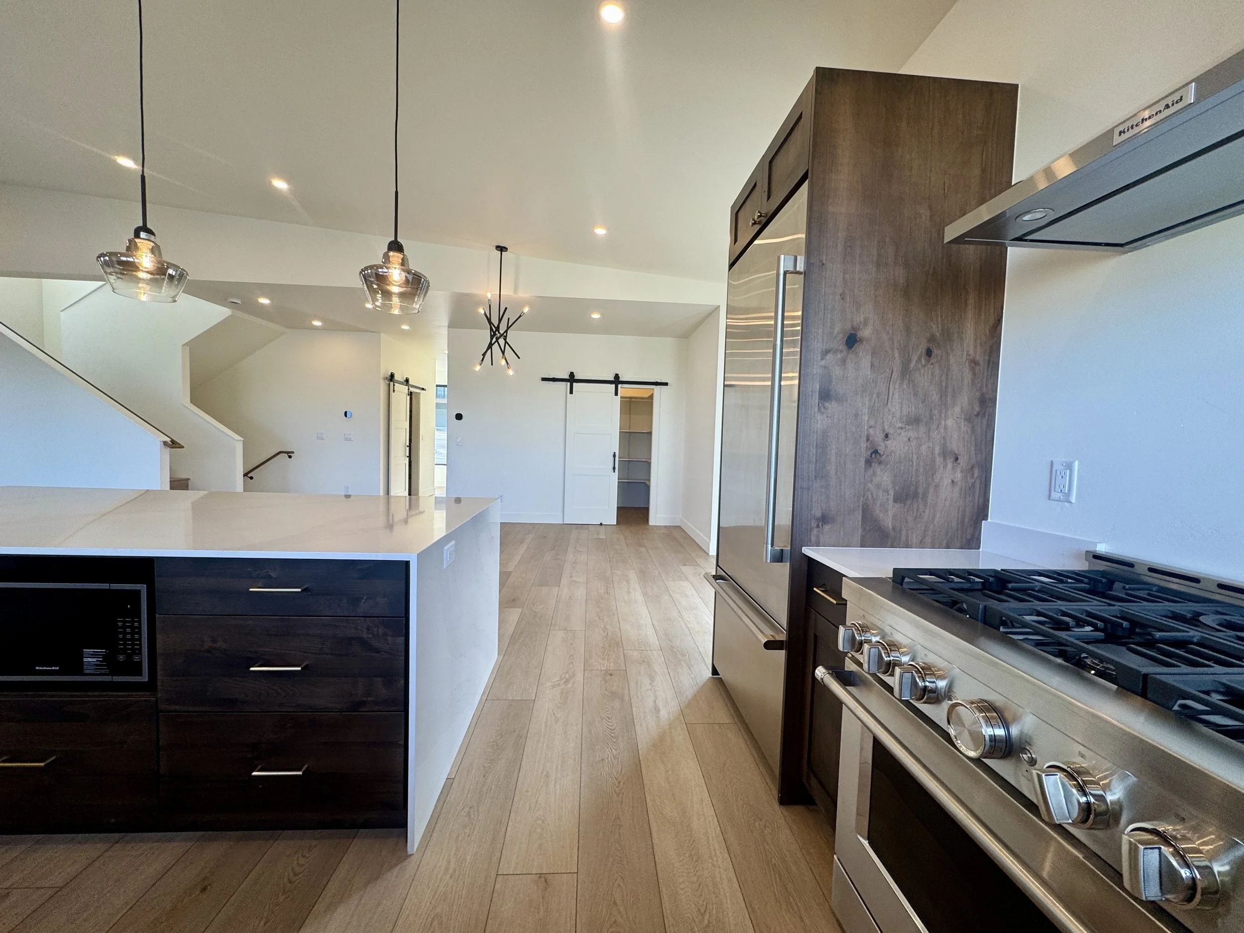 Modern kitchen with white island, dark wood cabinets, stainless steel refrigerator, gas stove, hardwood floors, hanging pendant lights, and sliding barn door in the background.