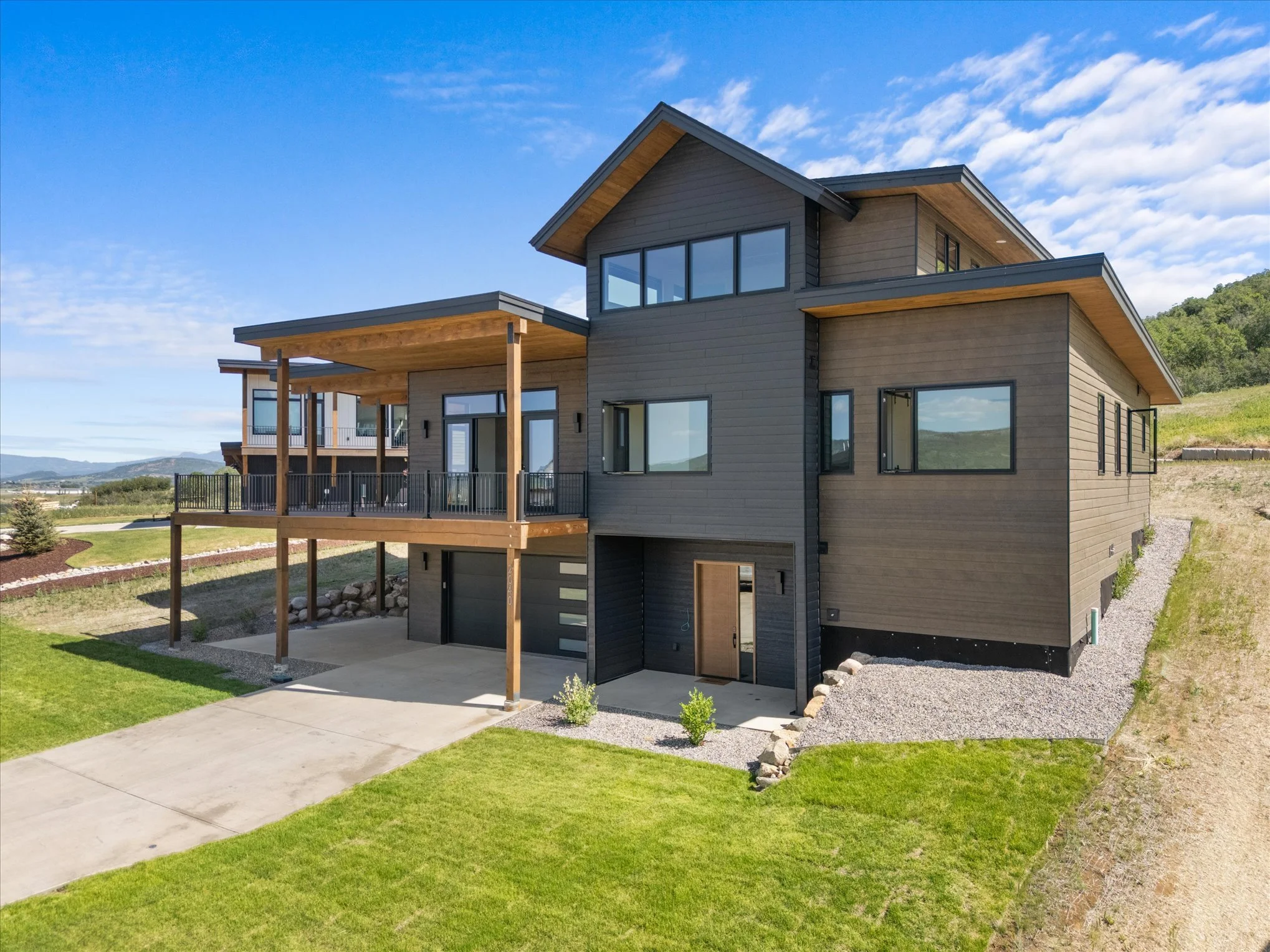 Modern three-story house with wood siding, large windows, and a spacious balcony with a black railing, situated on green lawns with a driveway and a backdrop of hills under a partly cloudy sky.