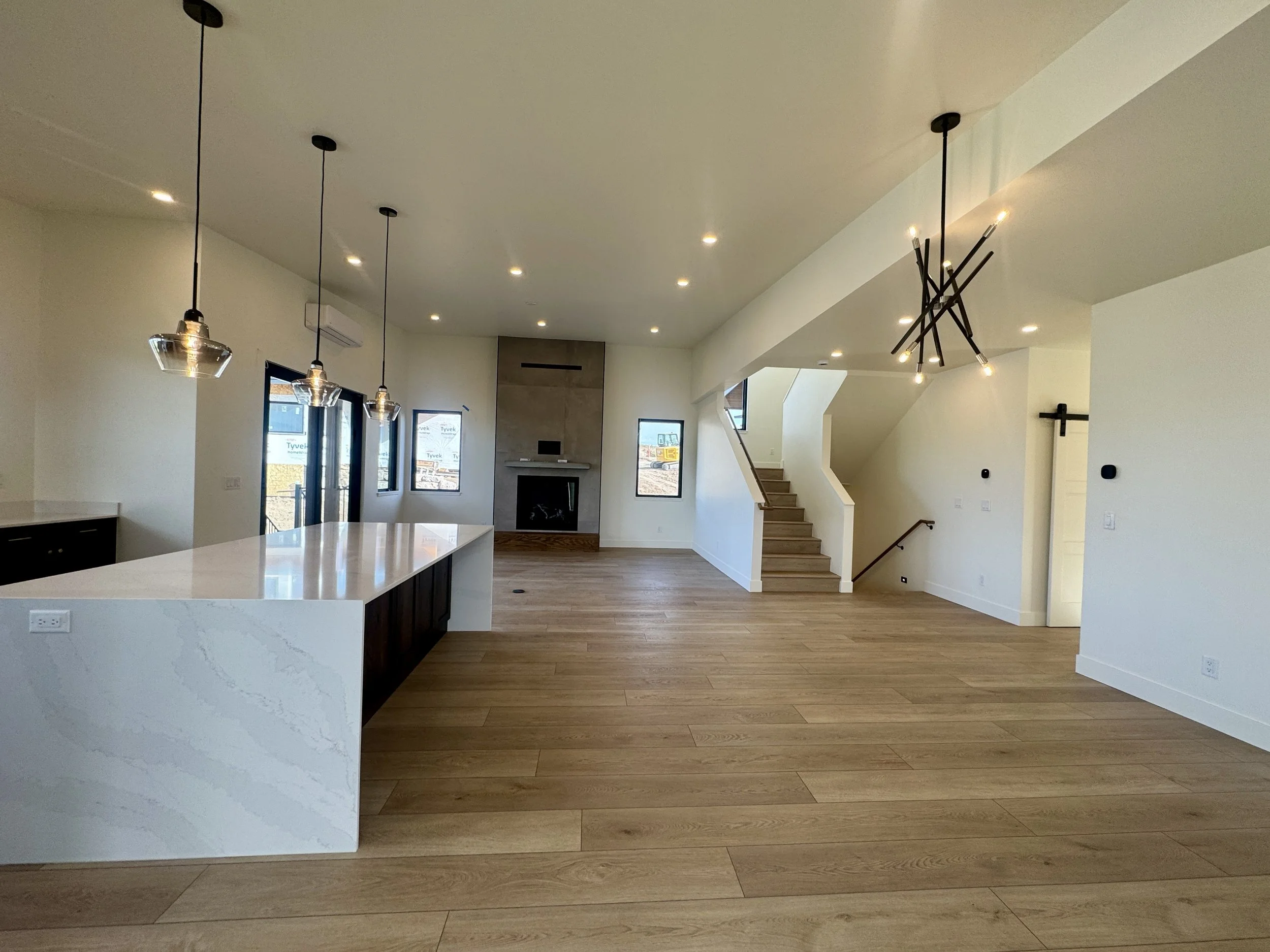 Spacious open-concept living area with wood flooring, modern lighting fixtures, a staircase leading upstairs, a fireplace, and large windows allowing natural light.