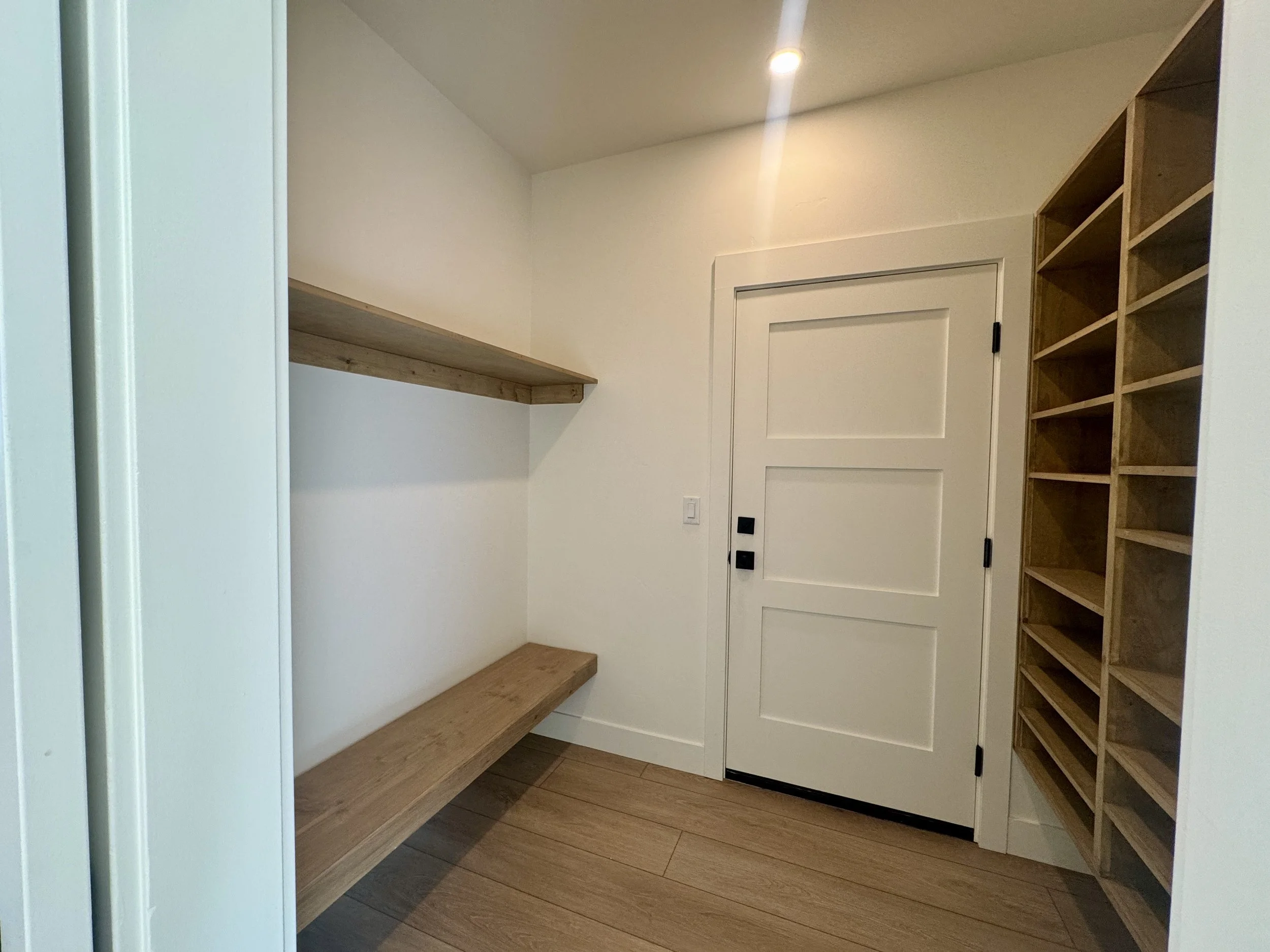 Room with wooden shelving on the right, a wooden bench on the left, a white door in the center, and a wooden shelf above the bench, with a small light fixture on the ceiling.