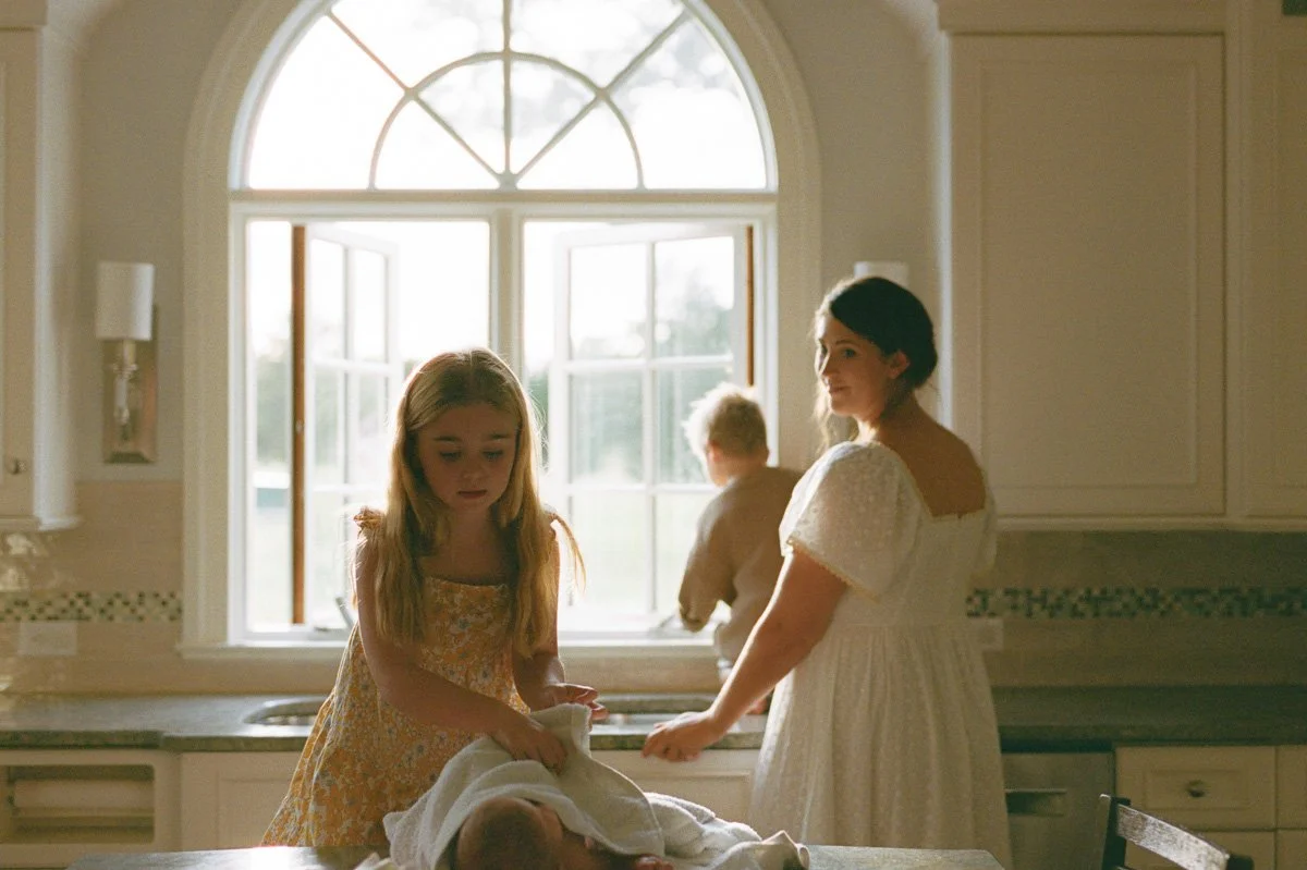 Connection and chaos during an in-home motherhood session with the Reich family in Greensboro. Mom looks on toward school-age daughter while she cares for her baby sibling after sink bath. Captured on 35mm film.