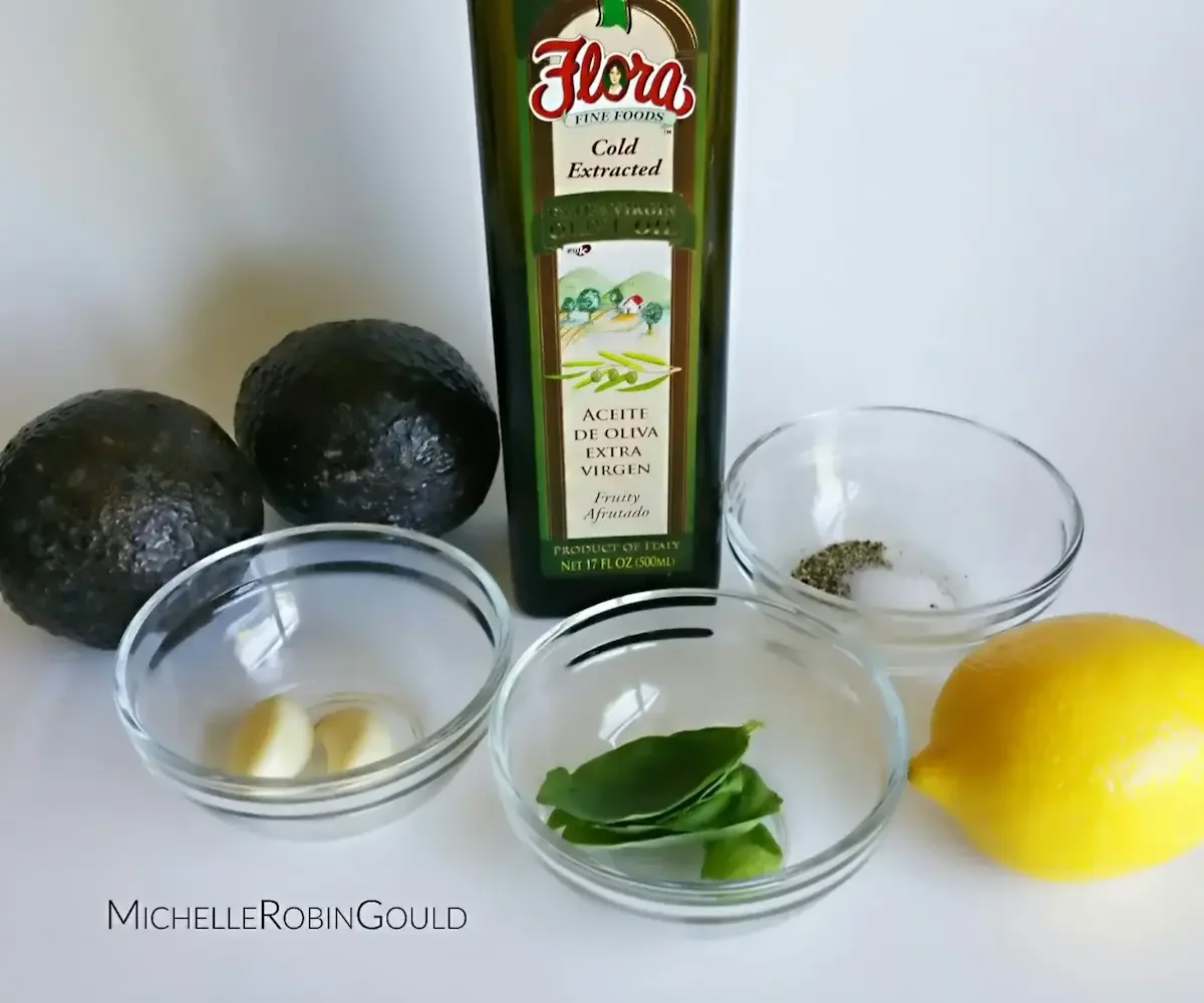 Two avocados, a bottle of extra virgin olive oil, a lemon, garlic cloves, basil leaves, and a bowl of salt and black pepper on a white surface, ingredients for a recipe by Michelle Robin Gould, EdD, LMHC.