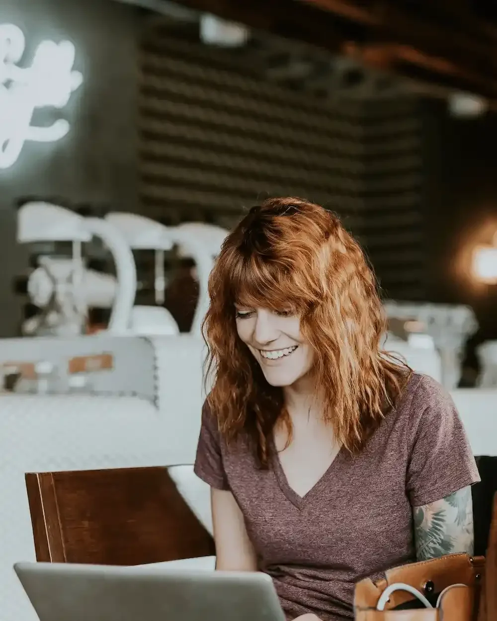 A casually dressed, tattooed young redheaded woman sits in a cafe talking to her online therapist on a laptop.