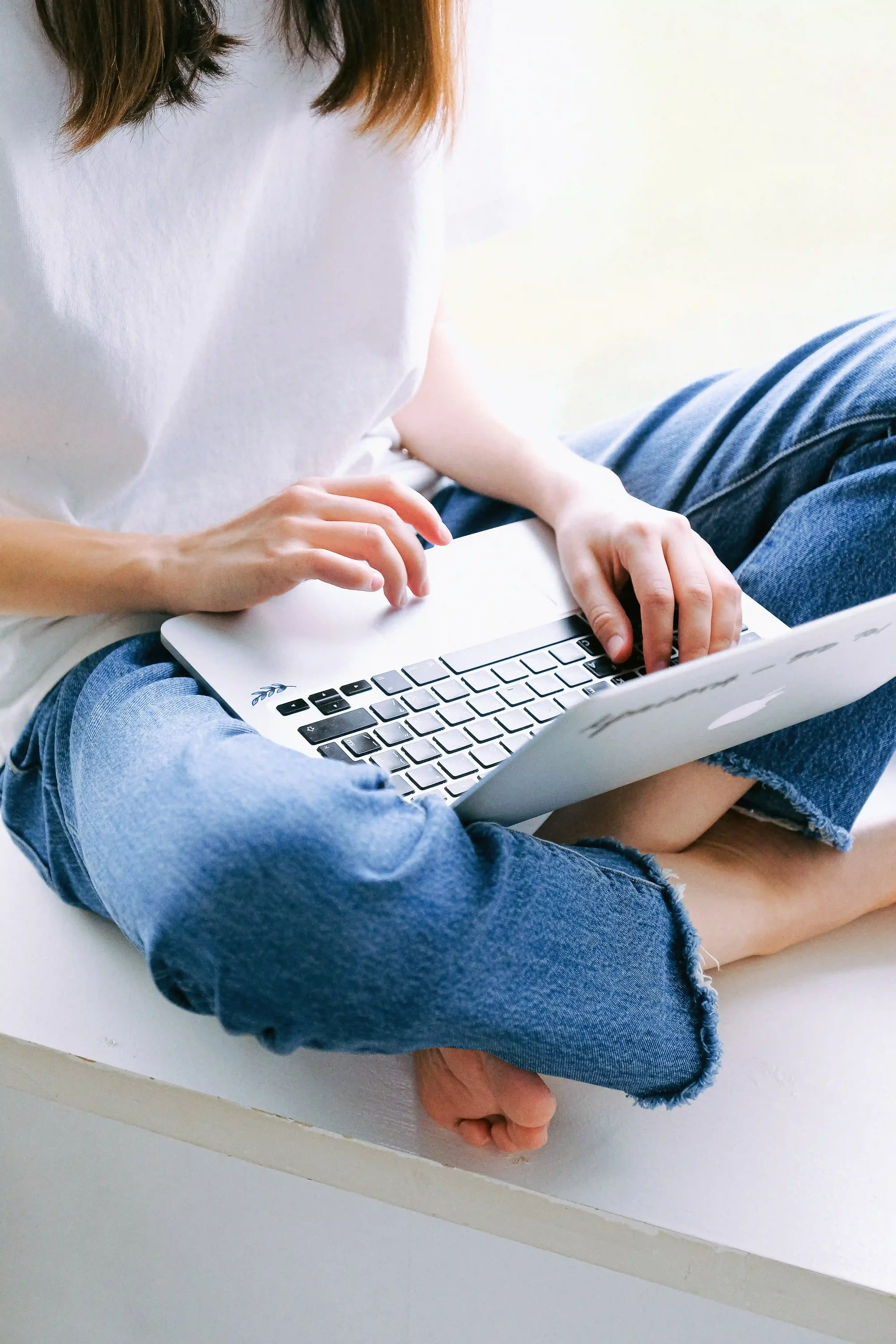 A woman in jeans and a t-shirt sits cross-legged with a computer on her lap as she signs in to start online therapy.