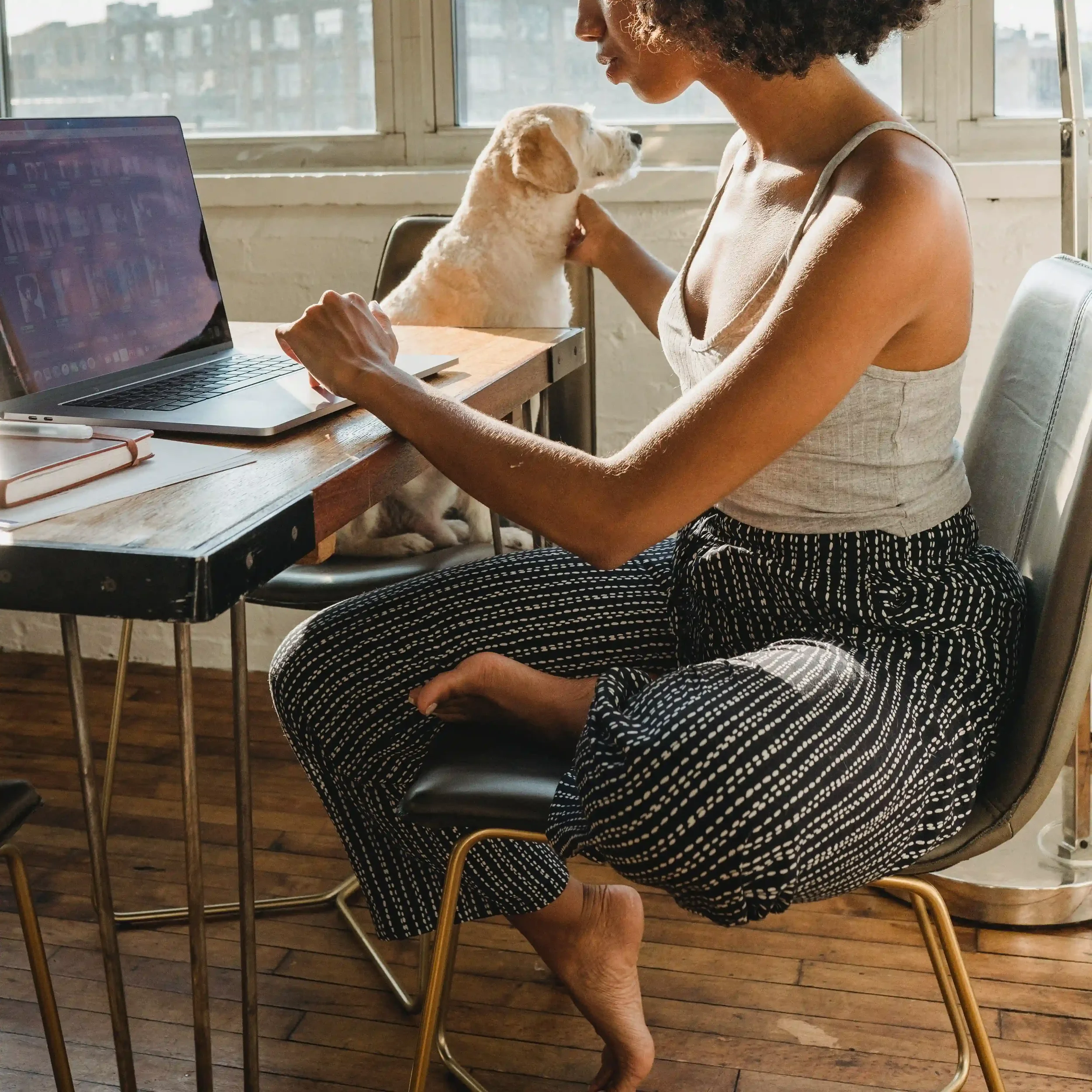 A young professional Black woman sits at the kitchen table with one hand on her dog and the other on her keyboard as she attends online therapy.