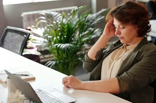 A professional woman sits at her work desk with her eyes closed, her hand raised to her temple, distracted by thoughts of shame and guilt due to addiction.