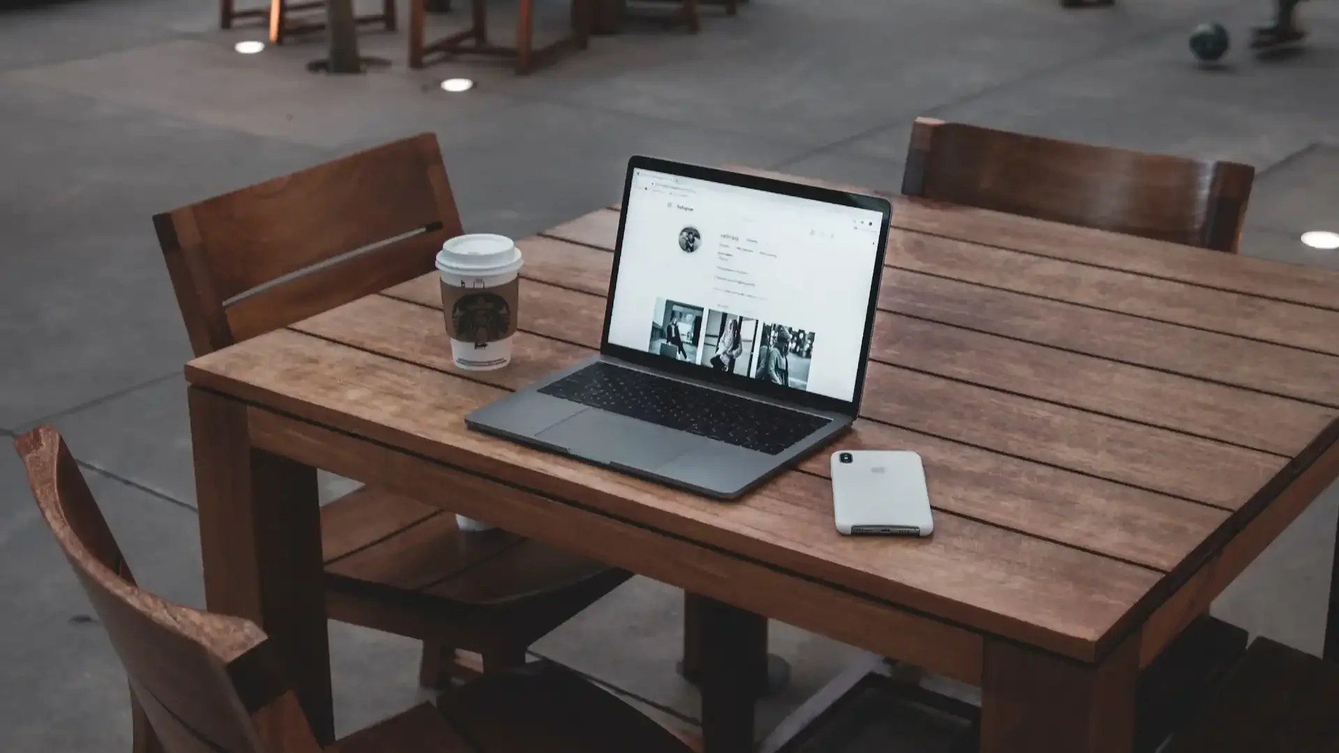 A laptop, cell phone, and a to-go cup of coffee sit on a wooden table outside a restaurant