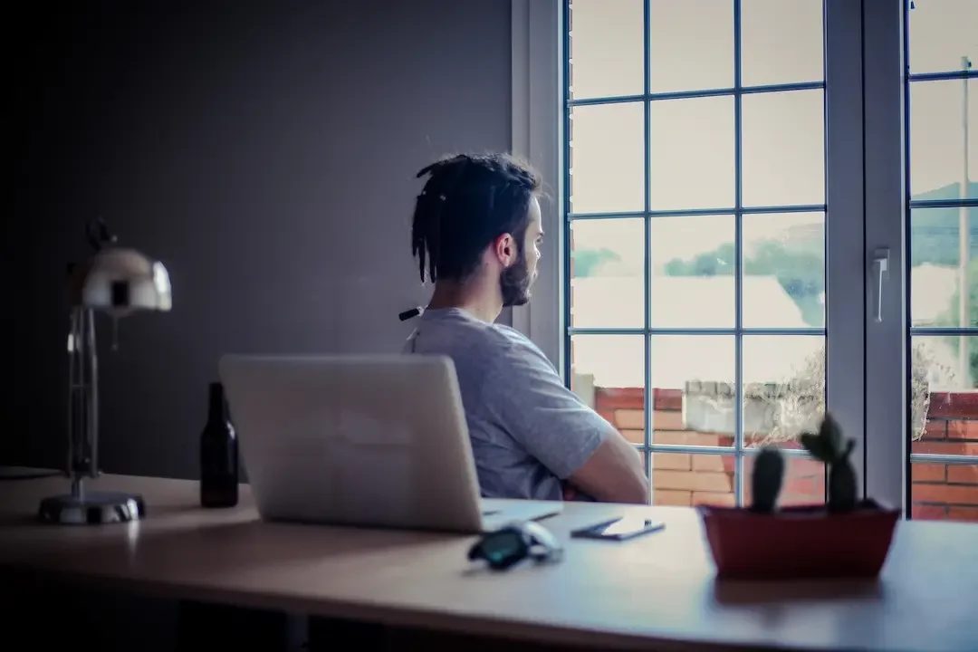 A young bearded man sits alone in his home office staring out the window feeling anxious. about his sobriety