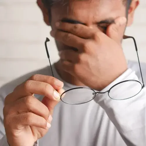 An adult man holds his glasses in one hand and rubs his eyes with the other hand, his eyes closed and his face tense while he feels trapped and powerless.