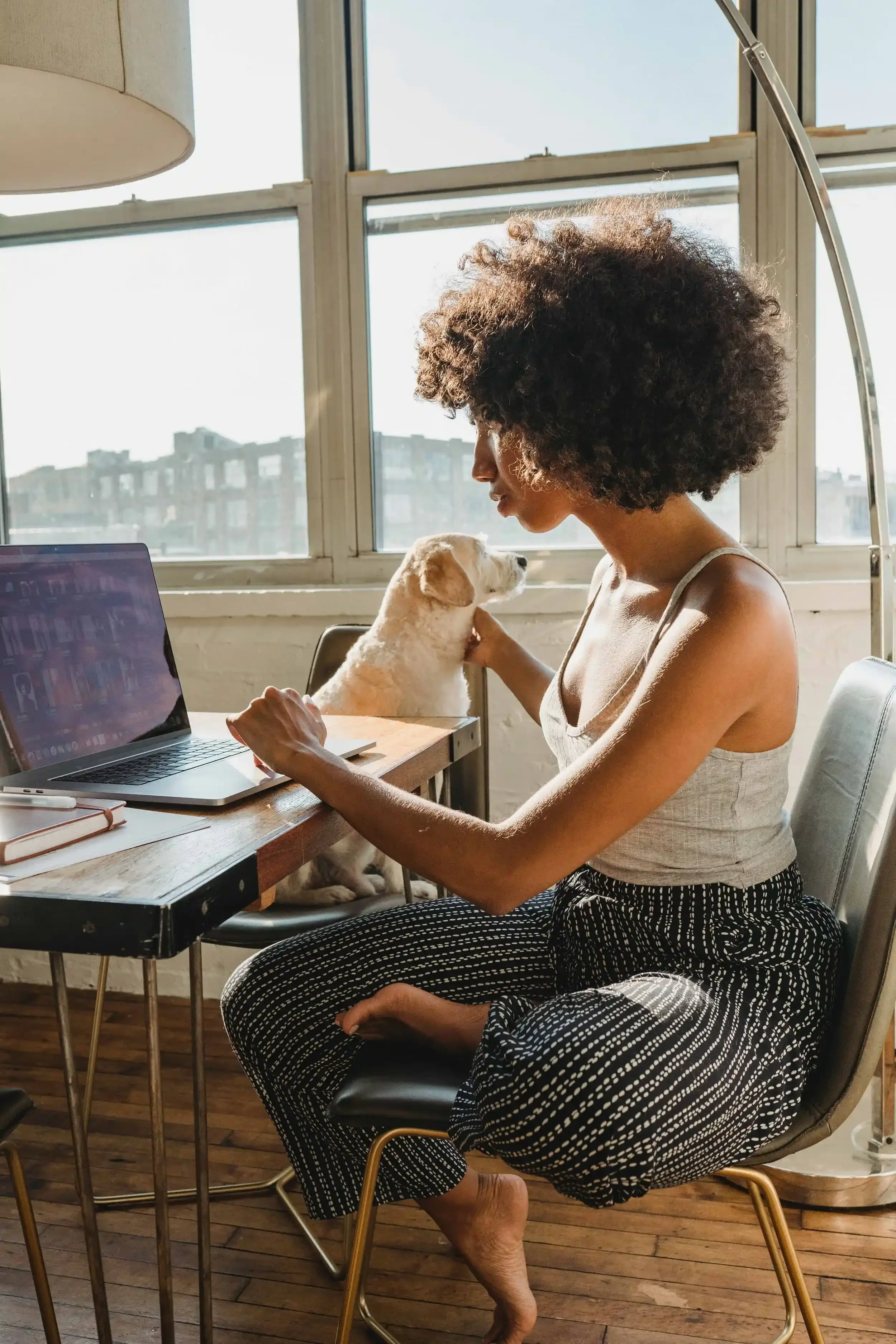 A casually-dressed Black woman logs onto her laptop on the kitchen table to attend online therapy with her dog at her side.