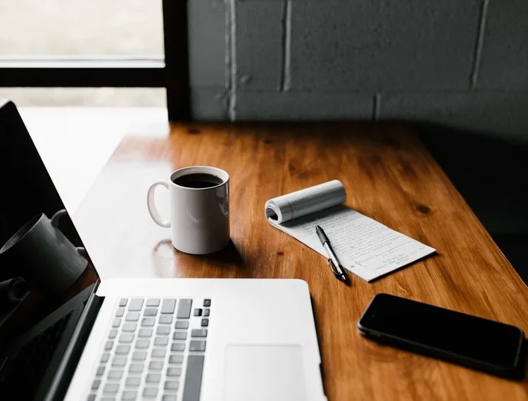 A laptop sits on a desk with a cup of coffee, a cell phone, and a note pad with pen