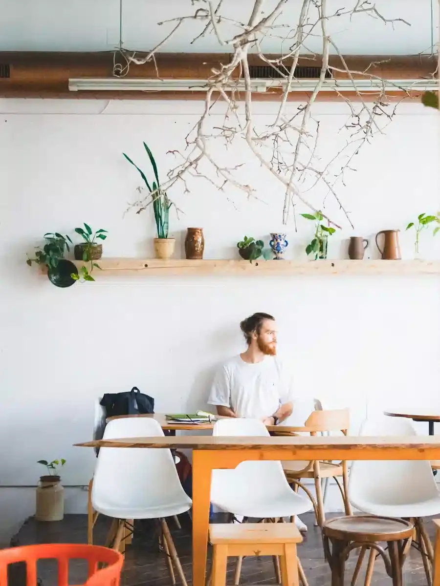 A man in his 30s sits in an open, airy cafe with books on the table and plants overhead as he looks out the window smiling.