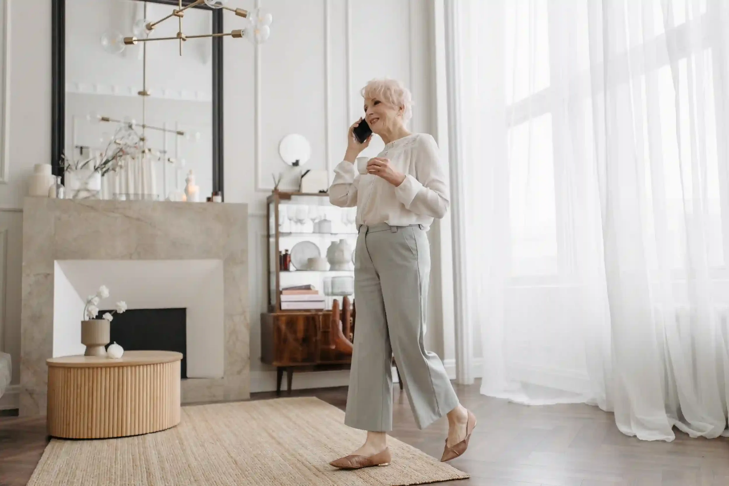 An elegant mature woman smiling while on a phone call in a bright, modern living room, conveying a sense of calm and personalized support as she talks to her concierge therapist in Florida..