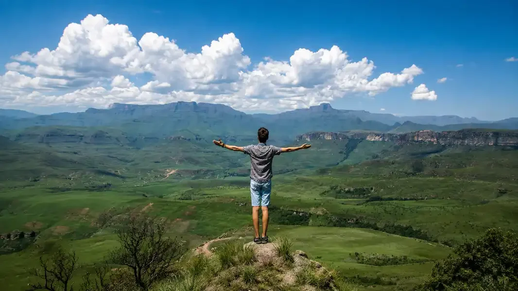 Man embracing addiction recovery with arms outstretched on a mountain overlook, overlooking a green valley and distant mountains under a bright blue sky.