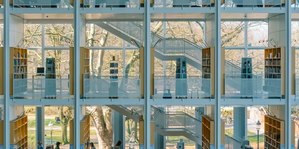 An interior view of a modern library featuring multi-level glass walls and white pillars, with bookshelves and people visible against a backdrop of trees outside.
