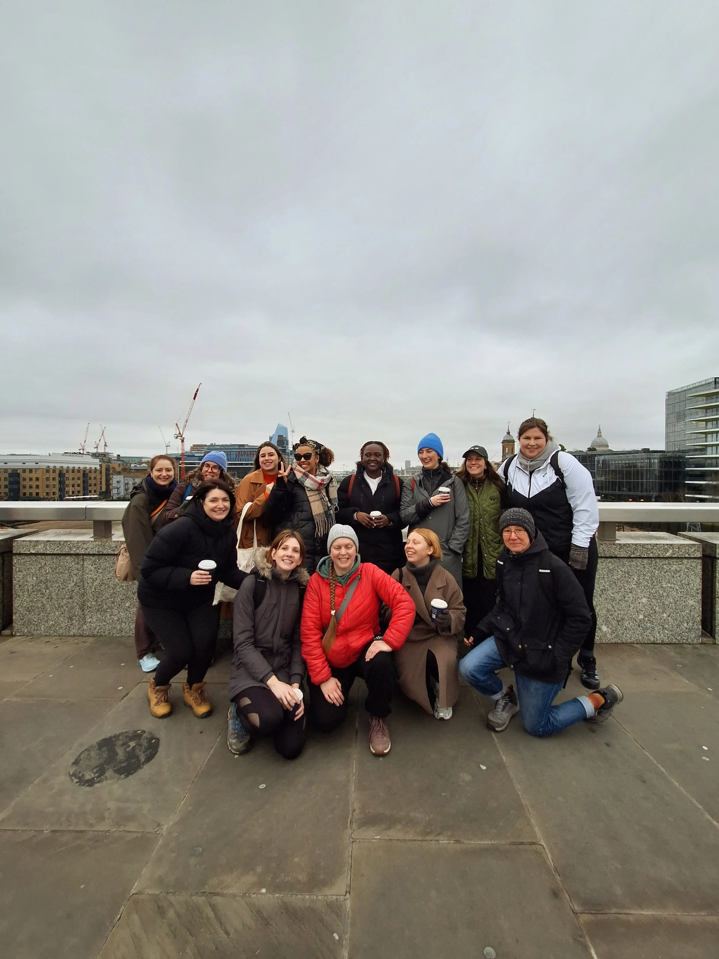 A group of smiling women, wearing coats and hats, kneeling and standing on an outdoor city rooftop with buildings and a cloudy sky in the background.