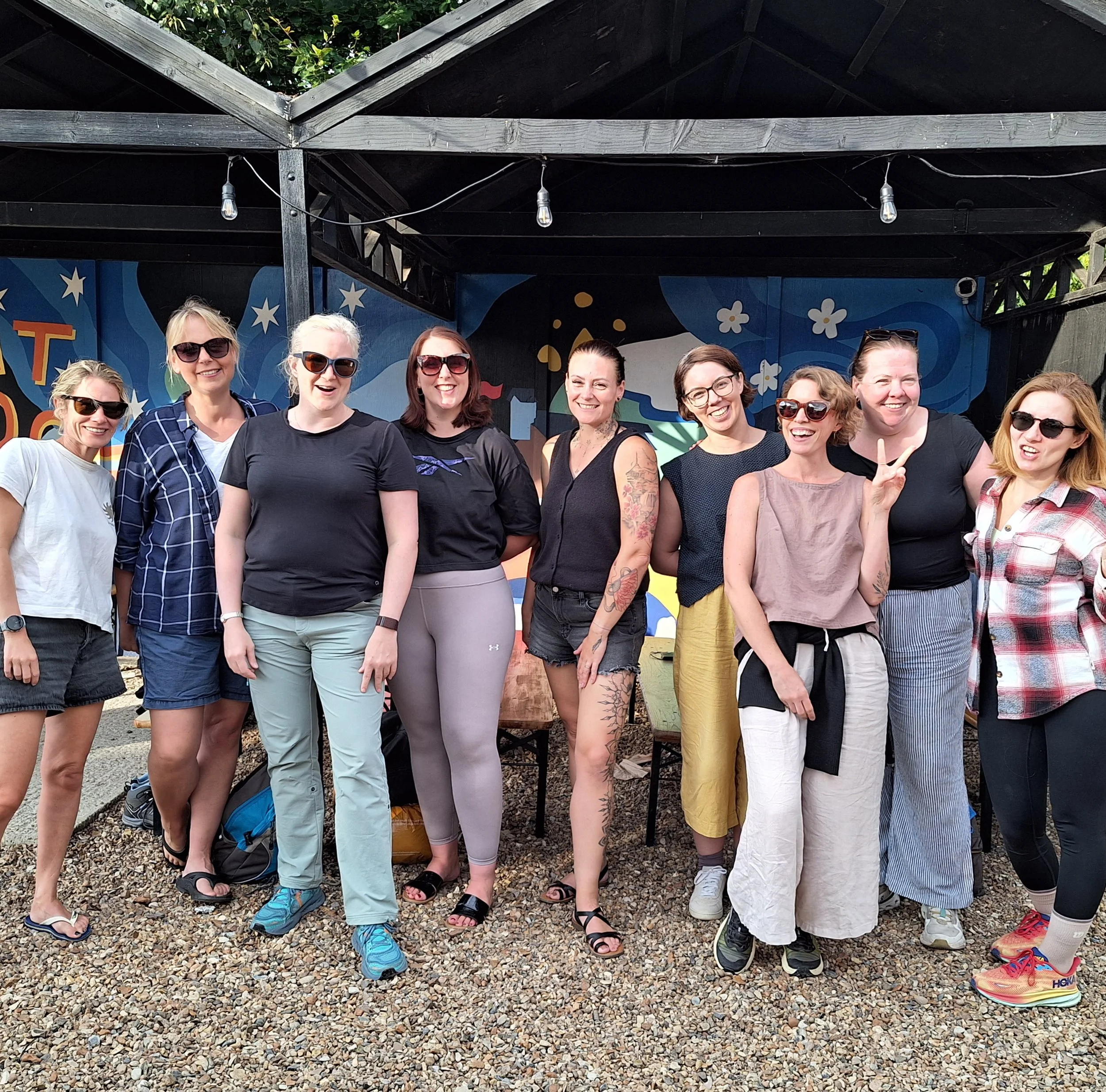 A group of women standing together outdoors on a gravel surface, smiling and posing for the photo under a black wooden pergola decorated with hanging string lights. The background features a colorful mural with stars and floral patterns.