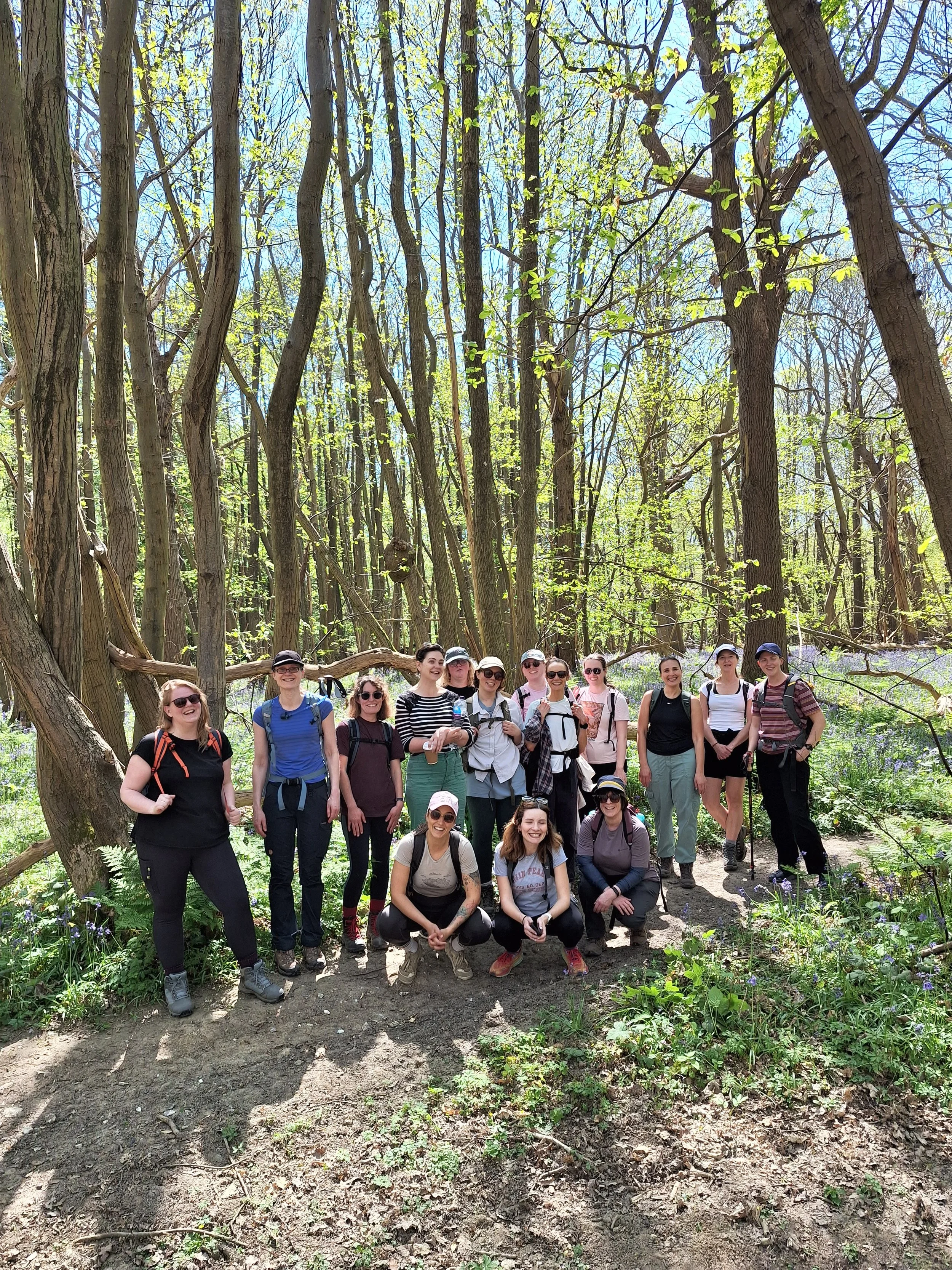 A group of smiling women standing together outdoors in a wooded area with tall trees and green foliage, some people are kneeling in front, during daytime.