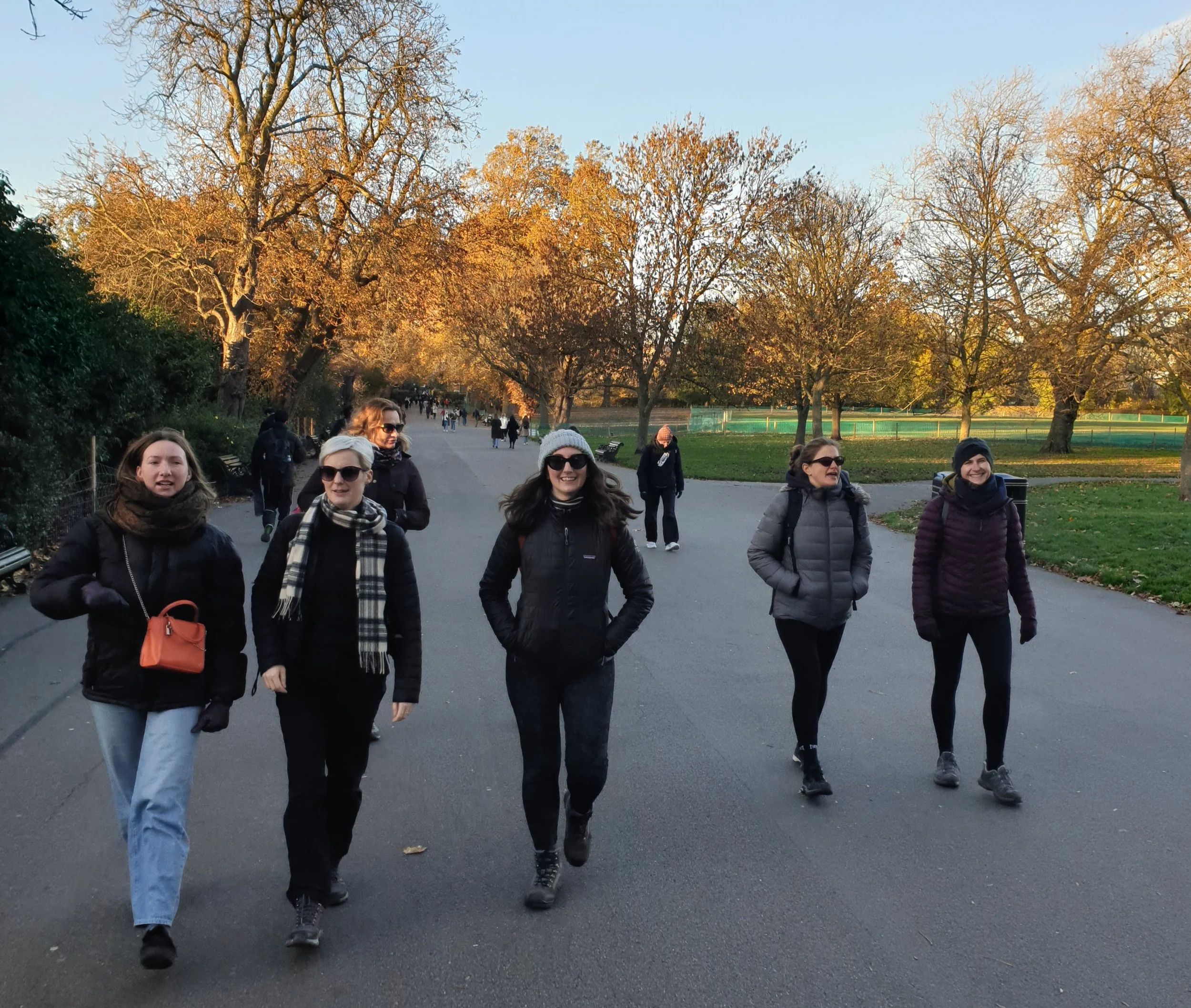 A group of women walking in a park on a sunny day during fall, with trees that have mostly bare branches and some leaves still on the trees.
