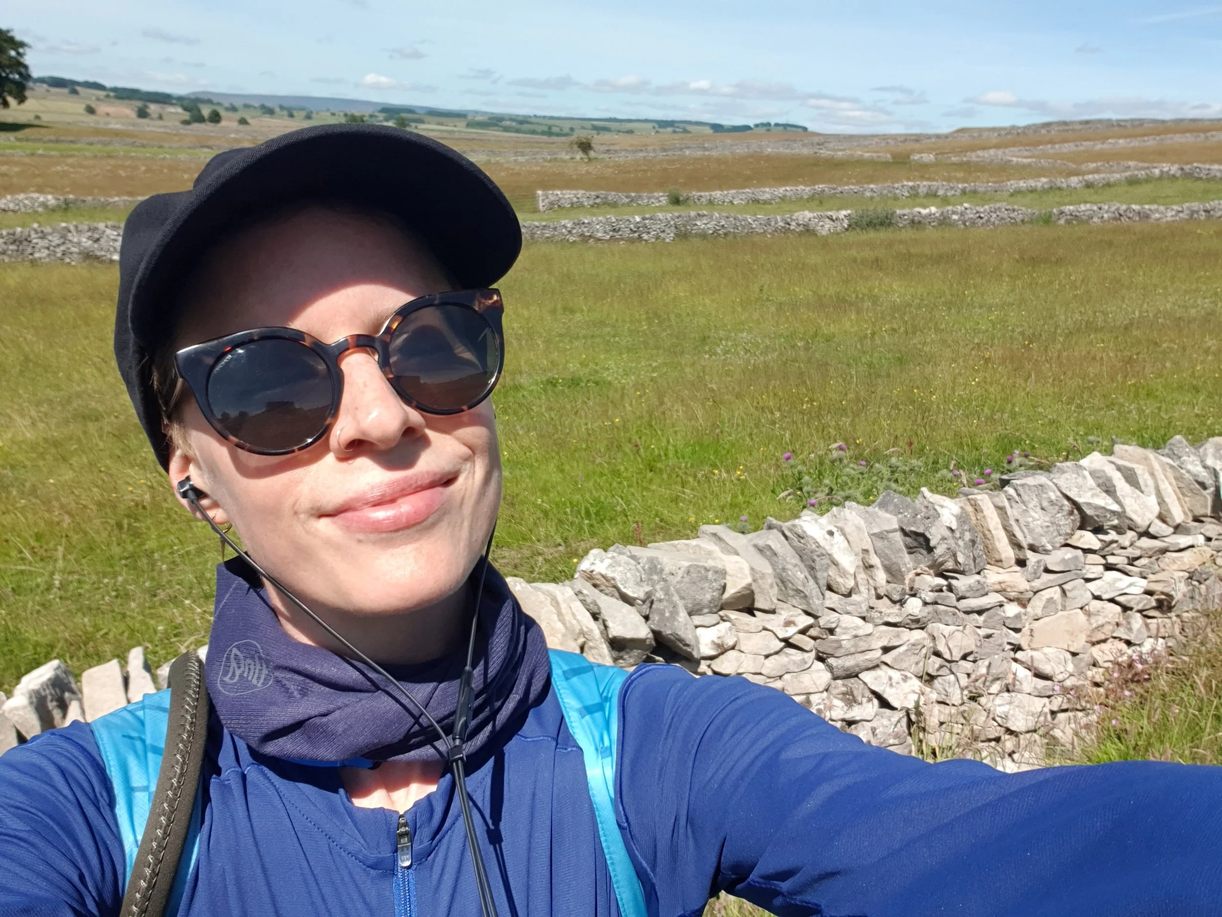 A smiling woman in sportswear, a cap and sunglasses, standing by a drystone wall with green fields behind her