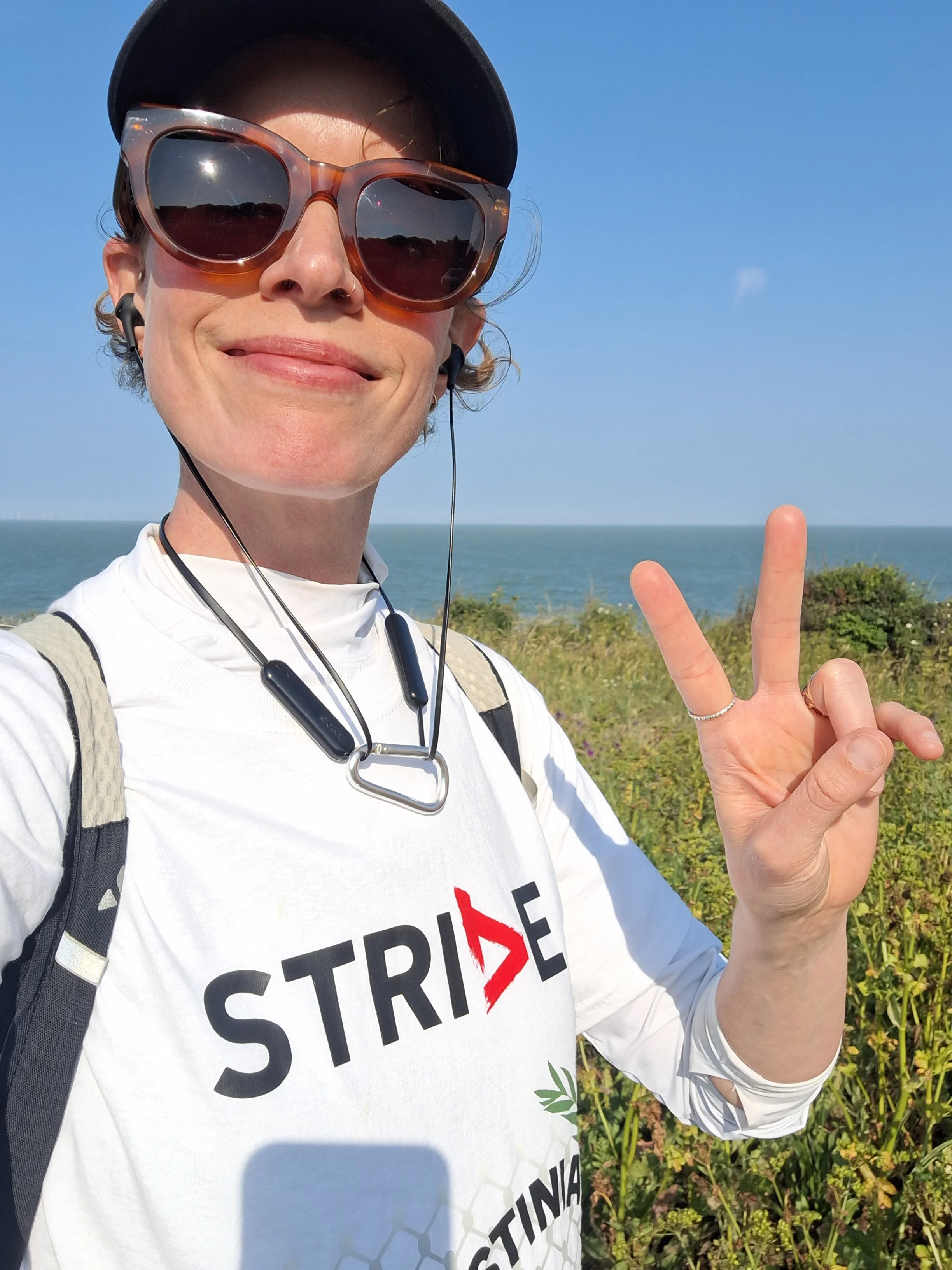 A woman taking a selfie wearing sportswear and a cap, smiling and holding up two fingers in a peace sign, standing in front of the sea