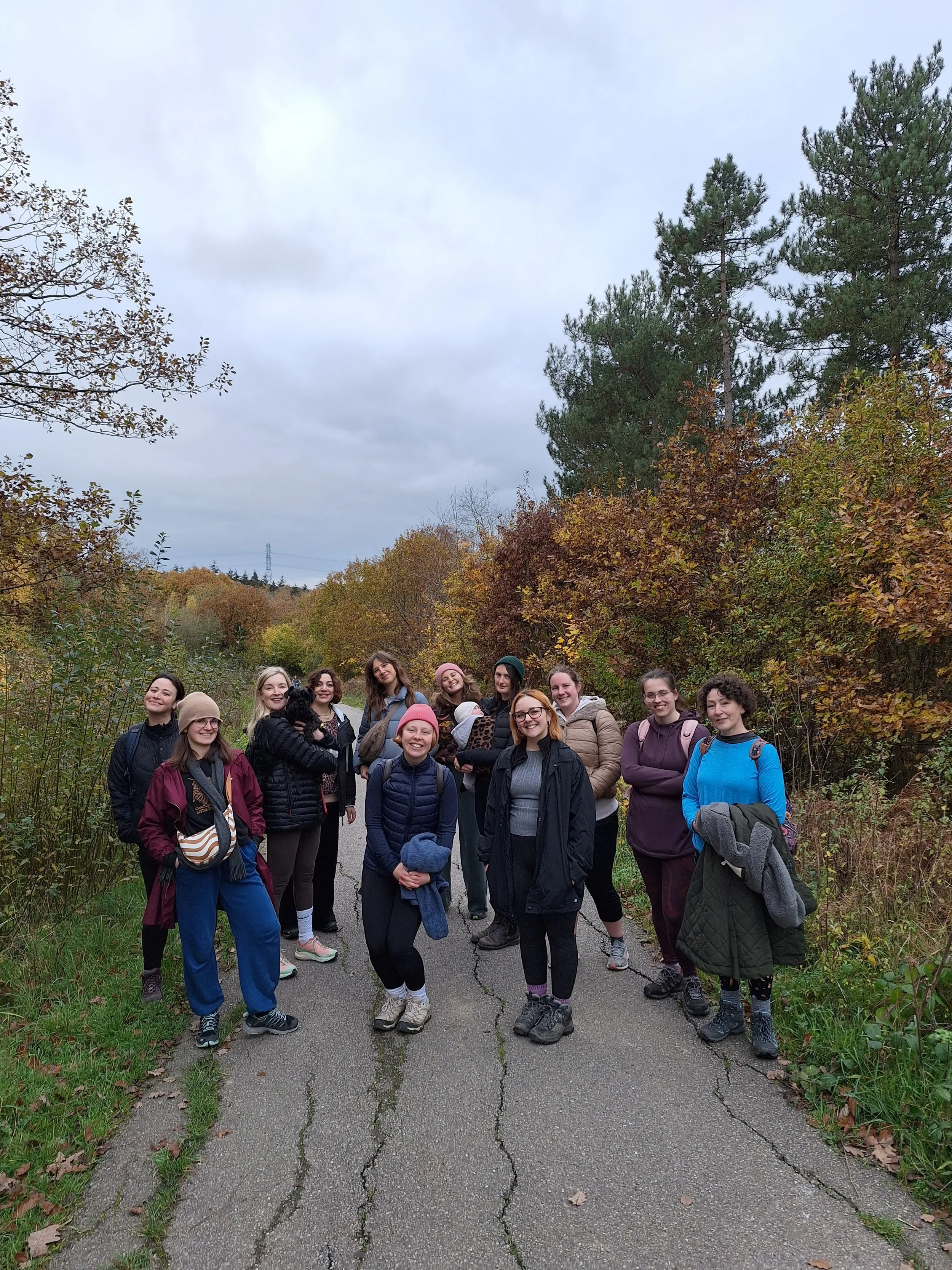 Group of women standing on a cracked paved trail surrounded by trees with fall foliage, overcast sky.