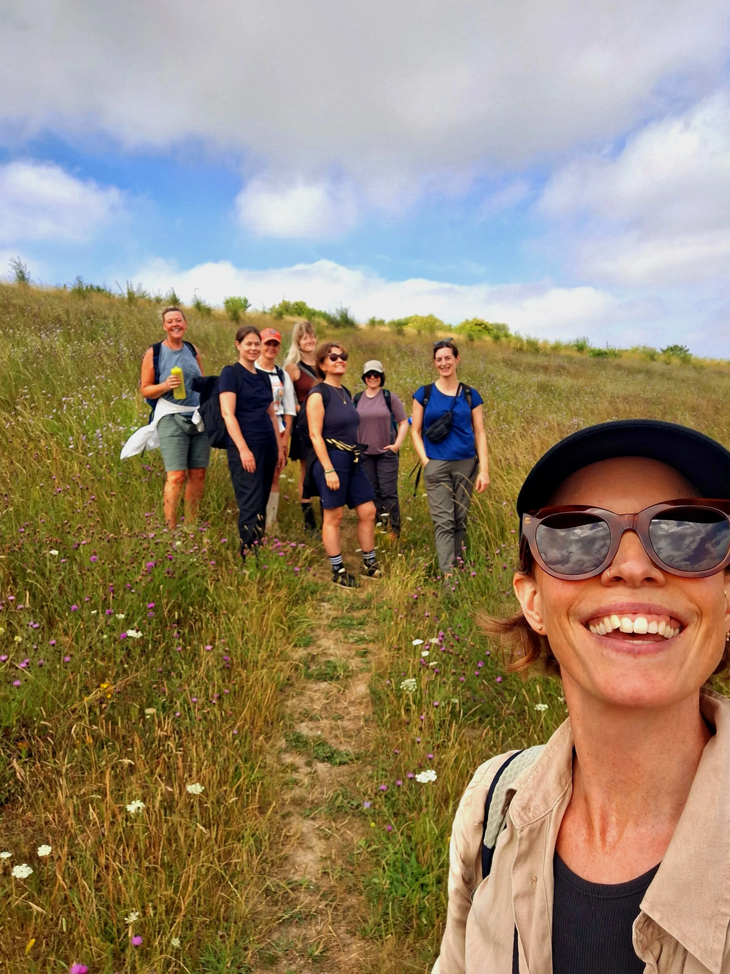 Woman smiling, taking selfie in front of seven other women, standing in a flower meadow