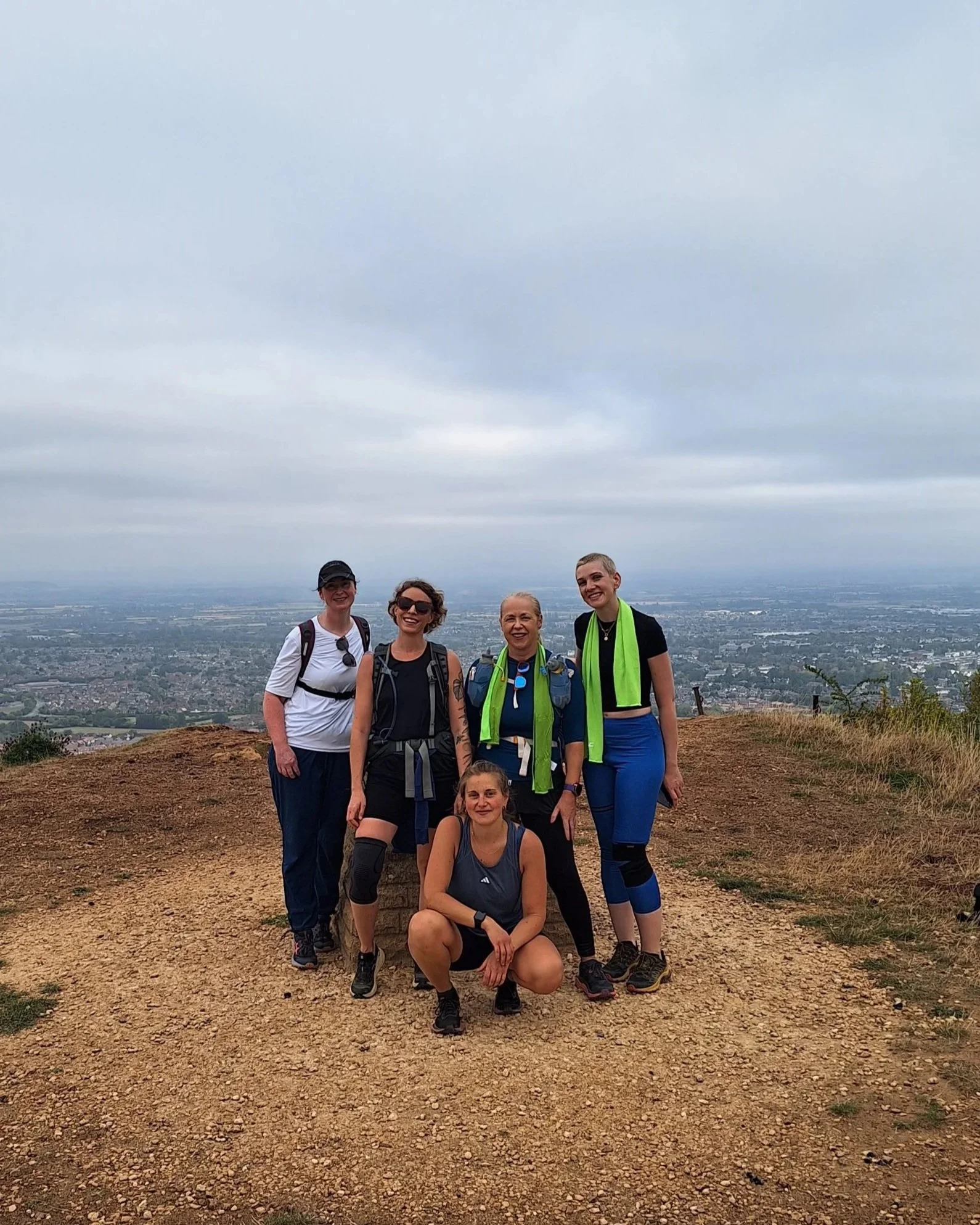 A group of seven women posing on a hilltop with a vast cityscape and cloudy sky in the background. They are wearing outdoor or hiking attire, some with backpacks and sunglasses.