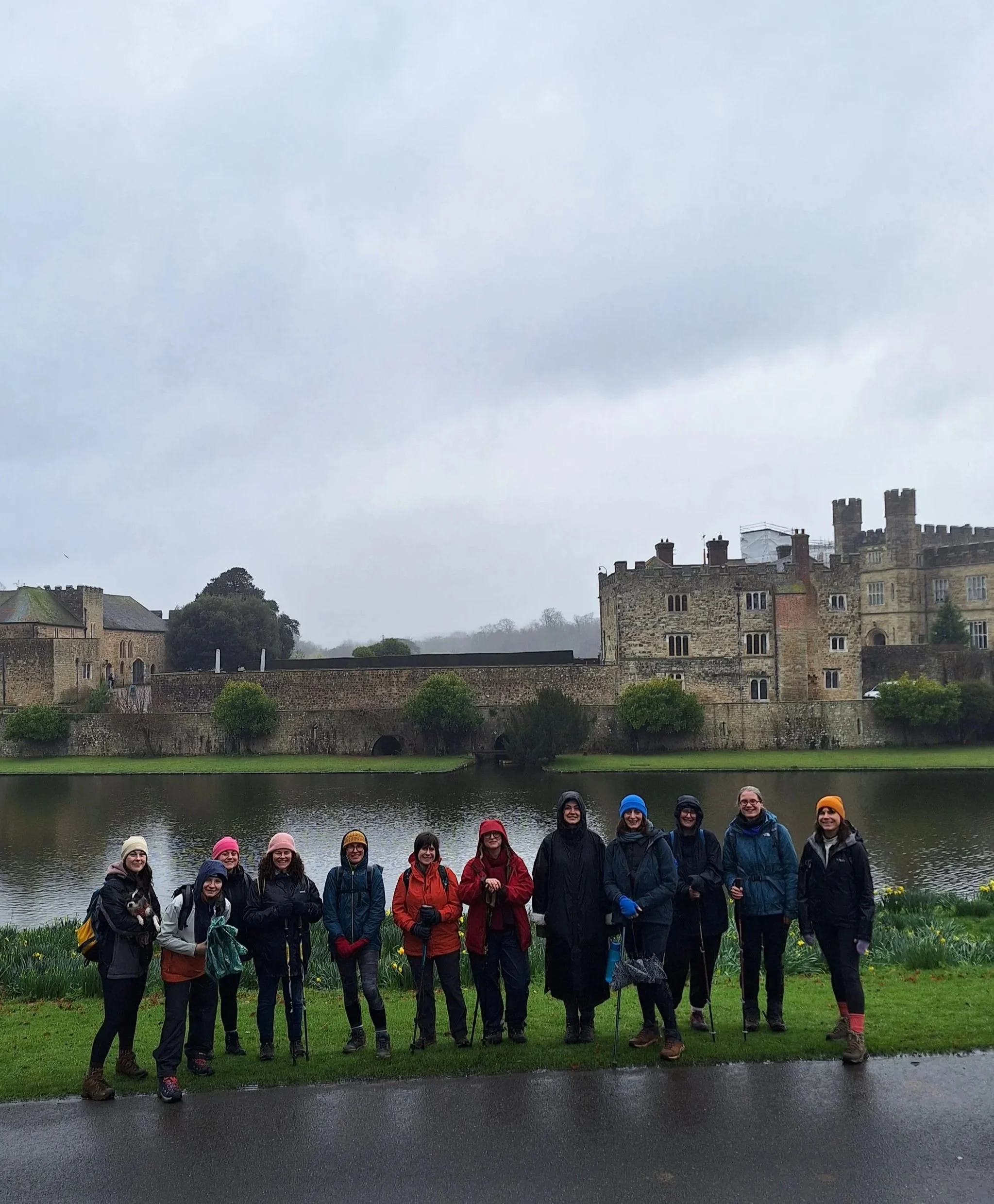 A group of women dressed in outdoor clothing standing together in front of a body of water, with historic castle buildings on a hill in the background on an overcast day.