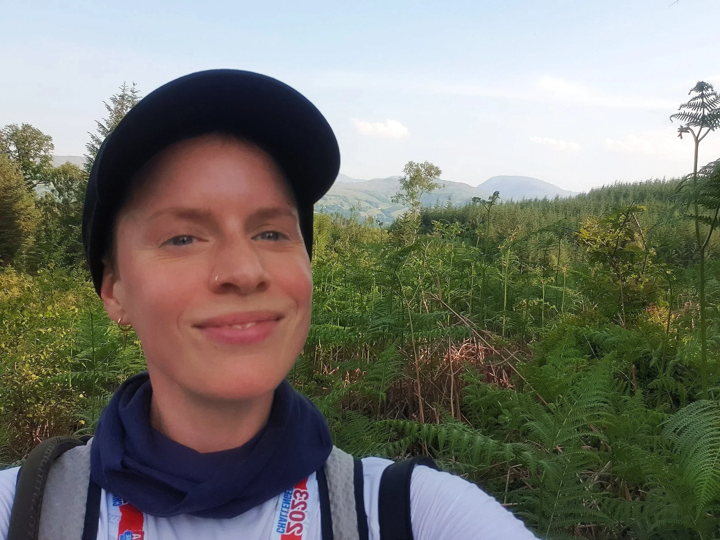 a smiling woman in a cap and sportswear standing in front of a view of lush greenery and mountains