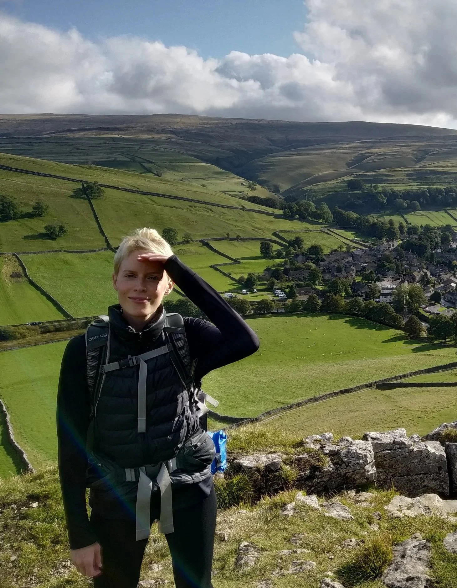 A woman standing on a hill with a view of a countryside valley behind her, shielding her face from the sun