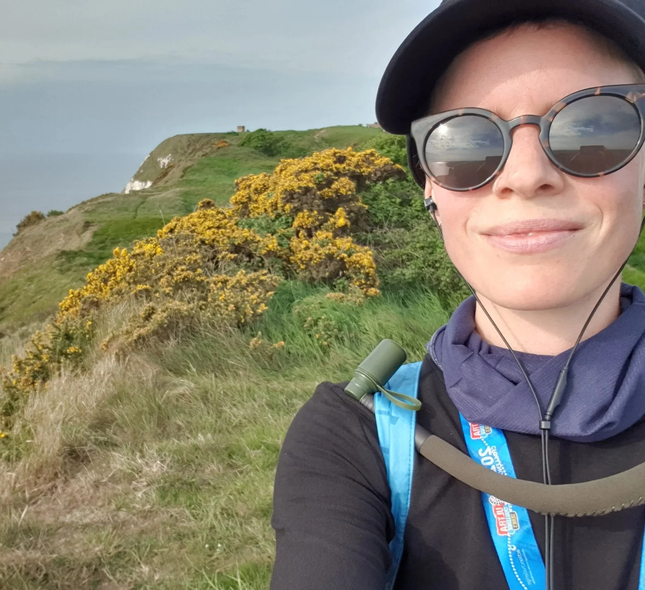 A woman in sportswear, a cap and sunglasses, standing on a clifftop smiling at the camera, with a view of white cliffs and greenery behind her