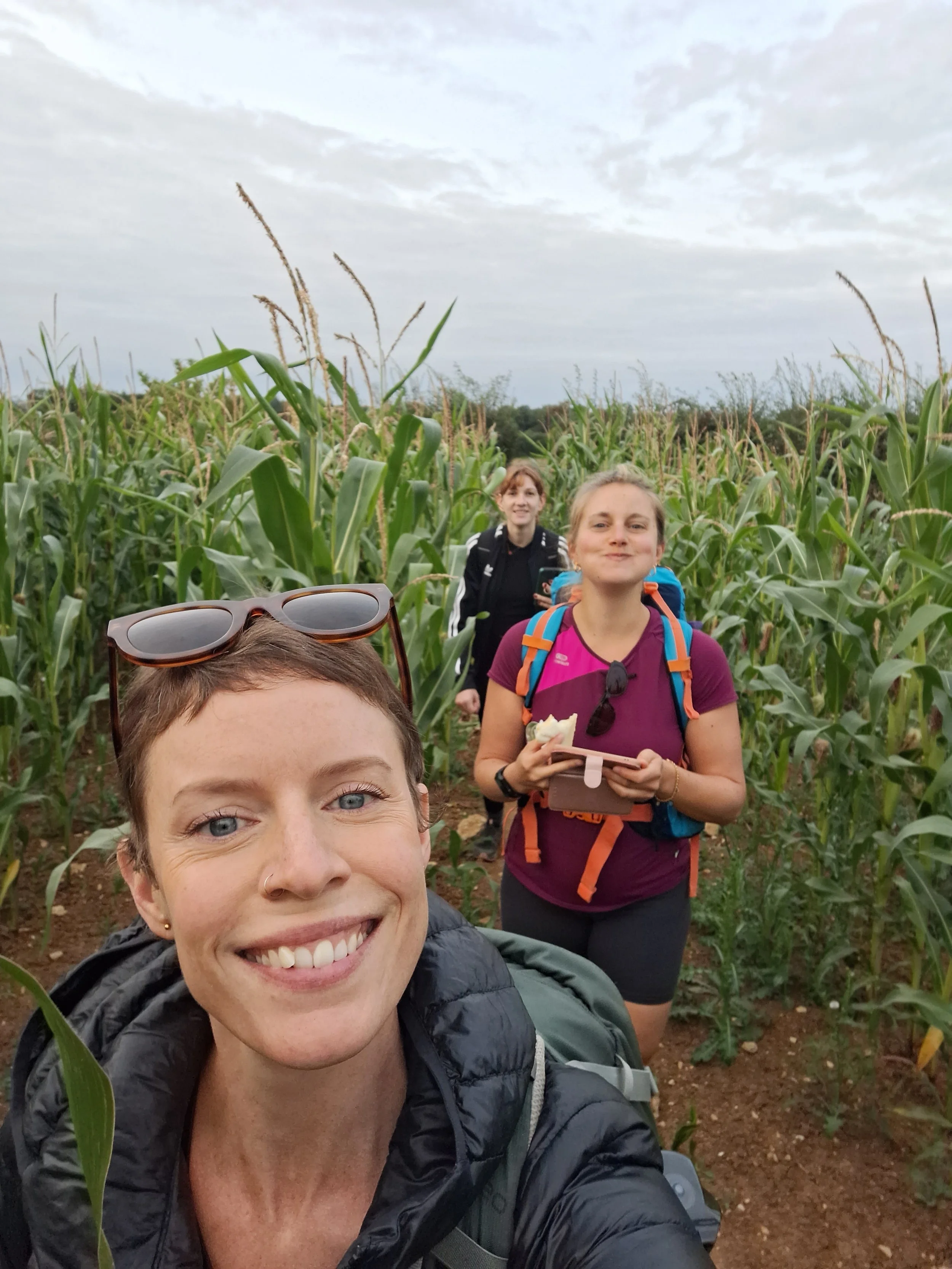 A woman taking a smiling selfie with two other women standing behind her, all wearing sportswear, in a filed full of tall corn plants