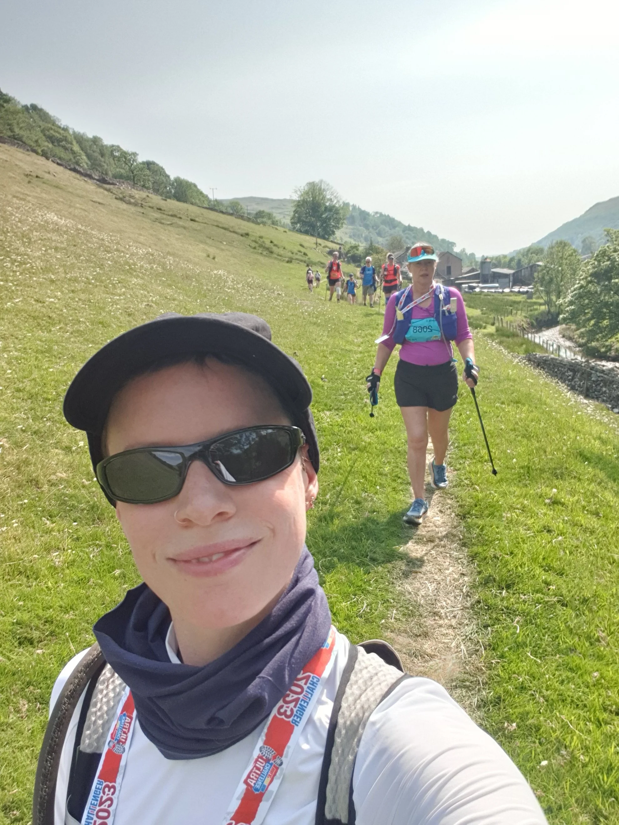 A woman taking a smiling selfie in sportswear, with several other people behind her in a countryside setting