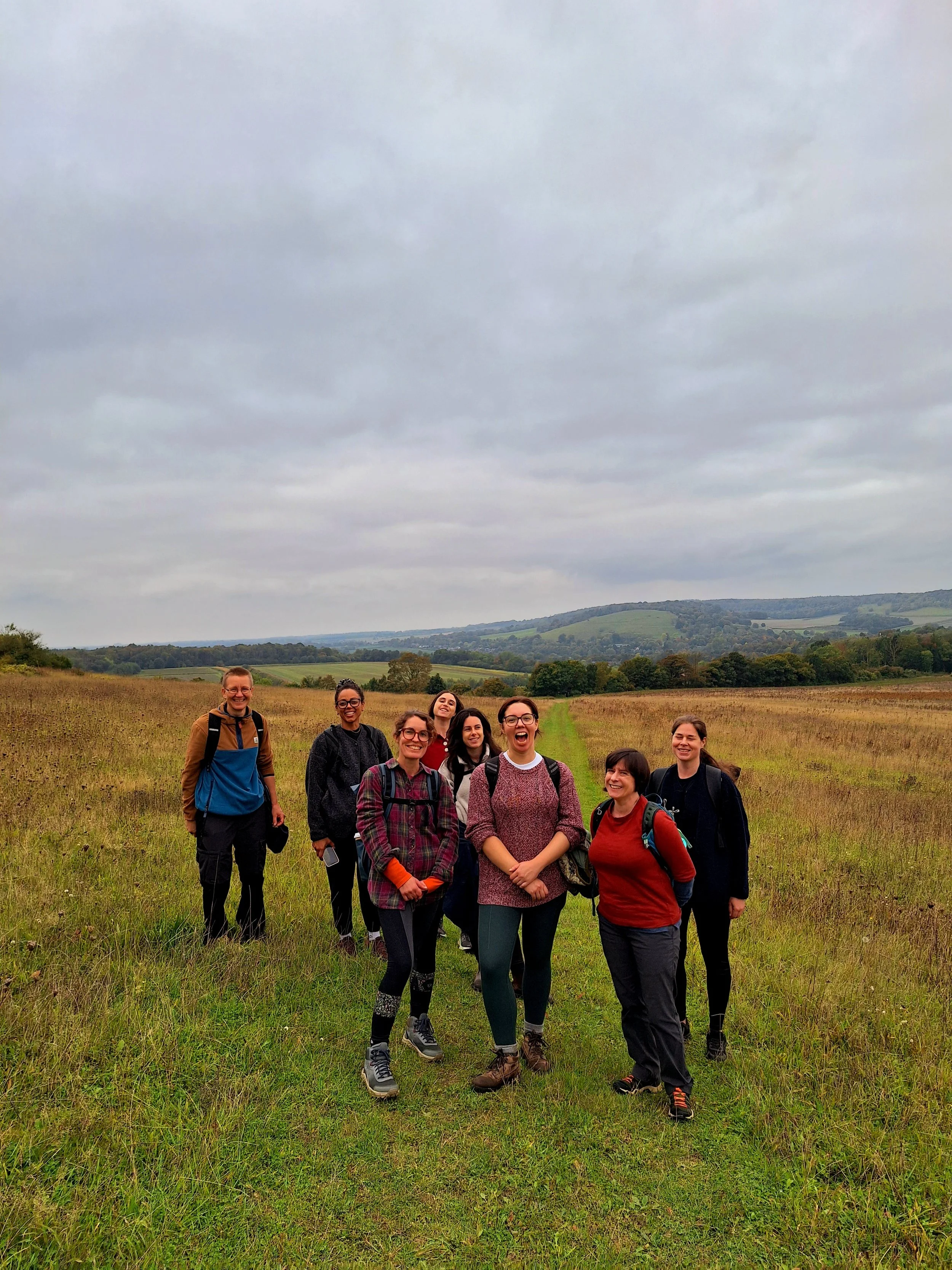 Group of eight women hiking along a trail through a grassy field with rolling hills and trees in the background under a cloudy sky.