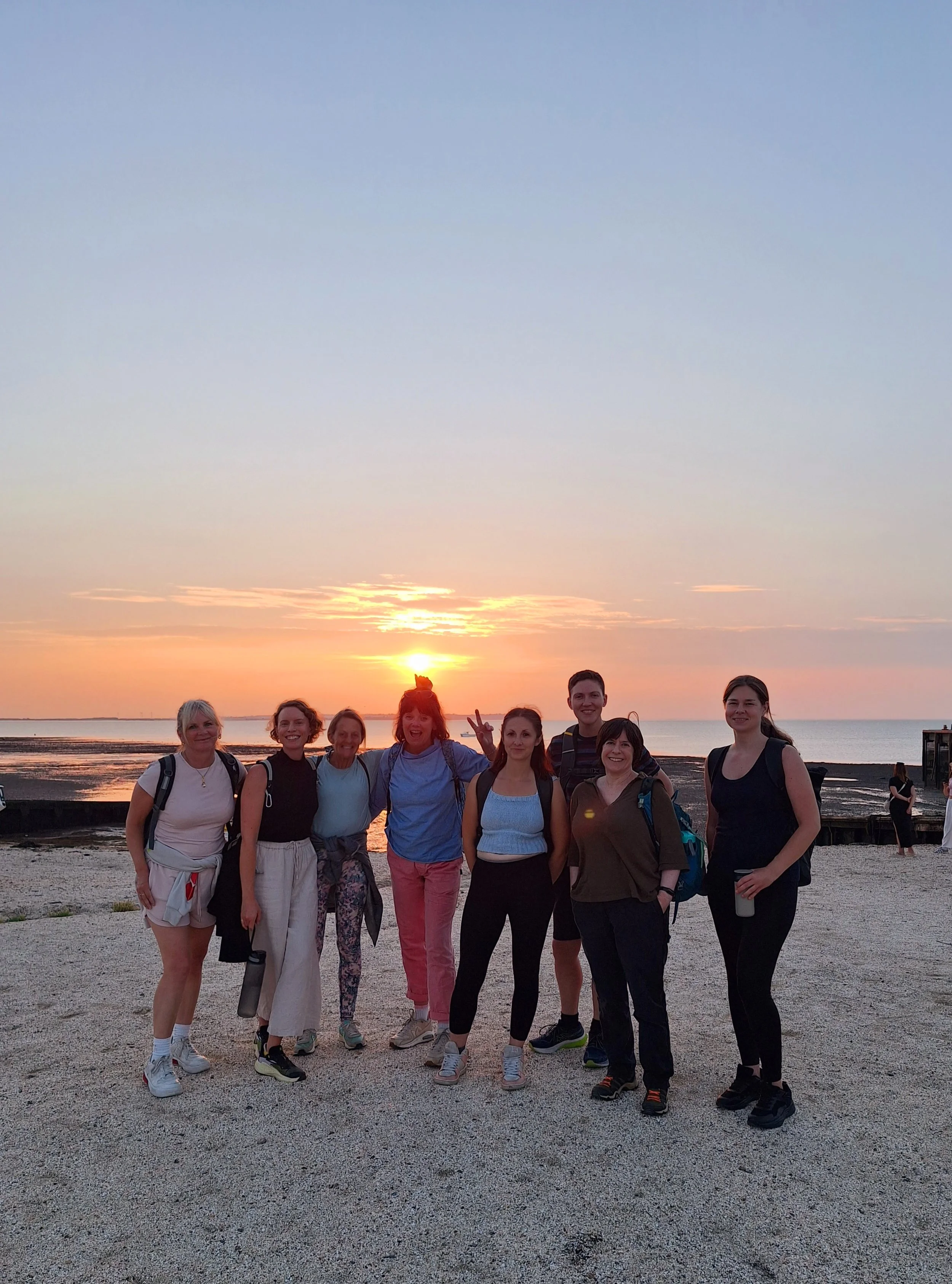 A group of nine women standing on a beach at sunset, smiling and posing for a photo.