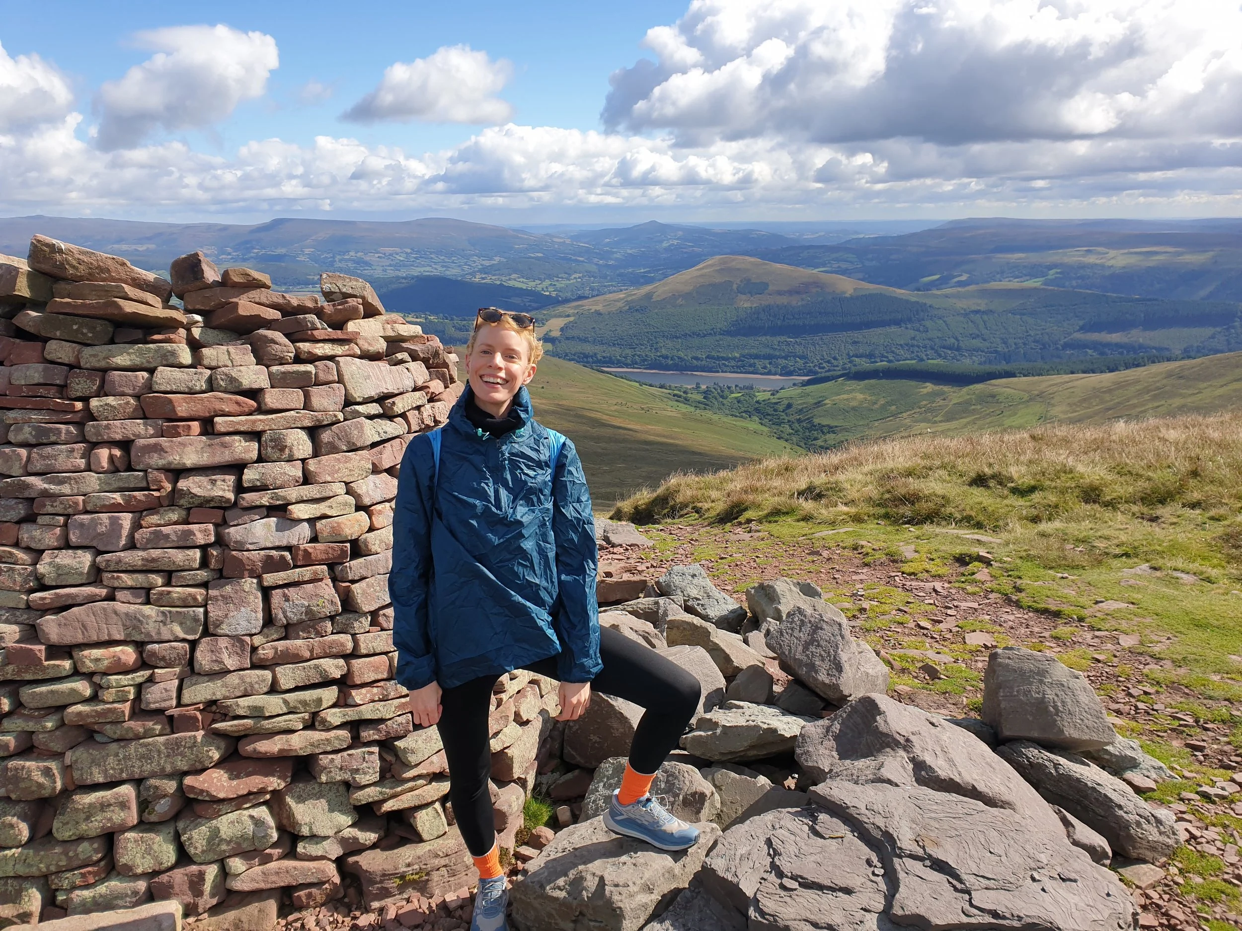 A woman in outdoorwear posing with one leg on a large rock and an old wall behind her, on top of a high hill with a sweeping view of hills and mountains behind her