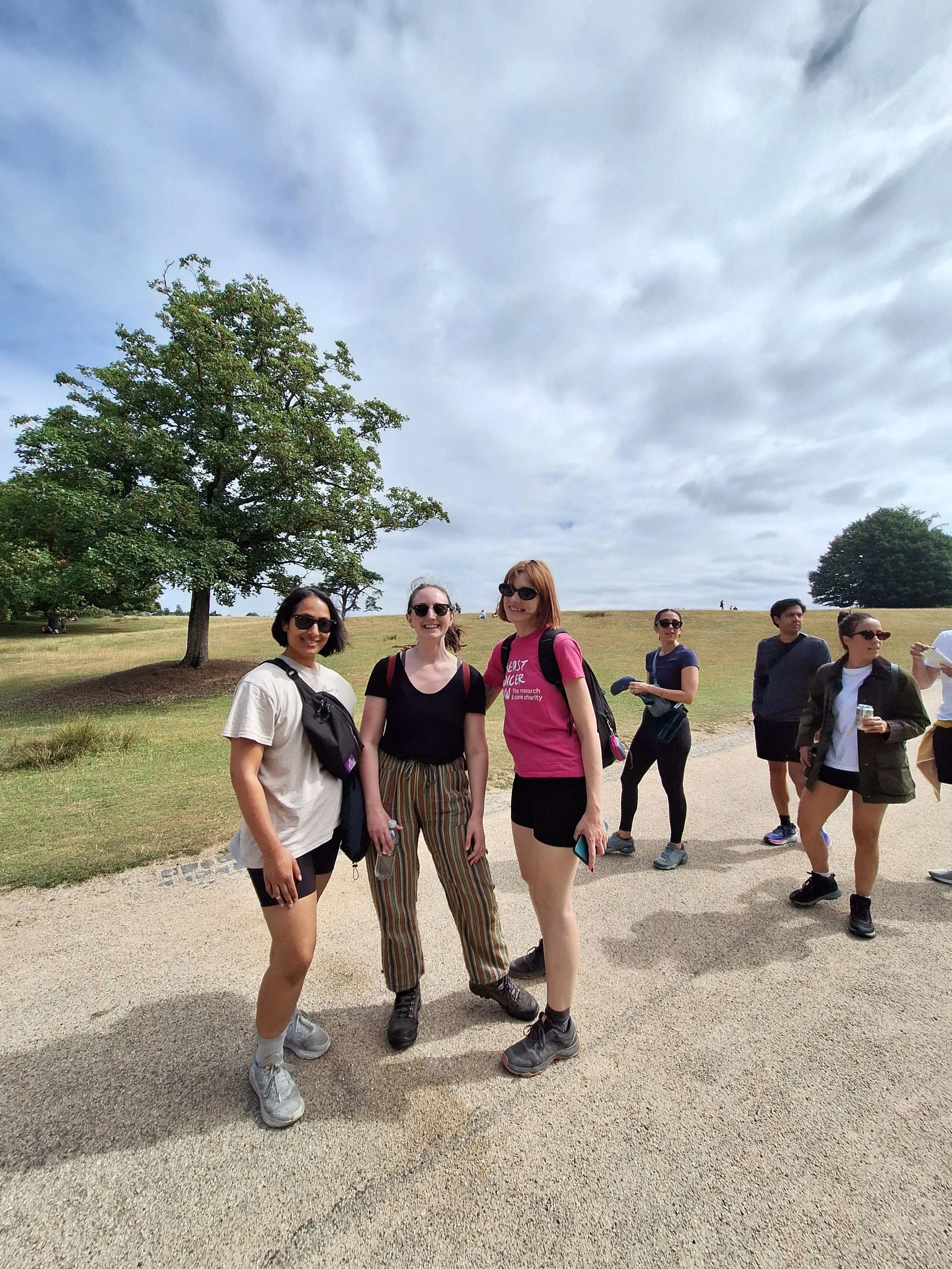 Group of women outdoors on a grassy area with a large tree and cloudy sky, some people smiling and walking.