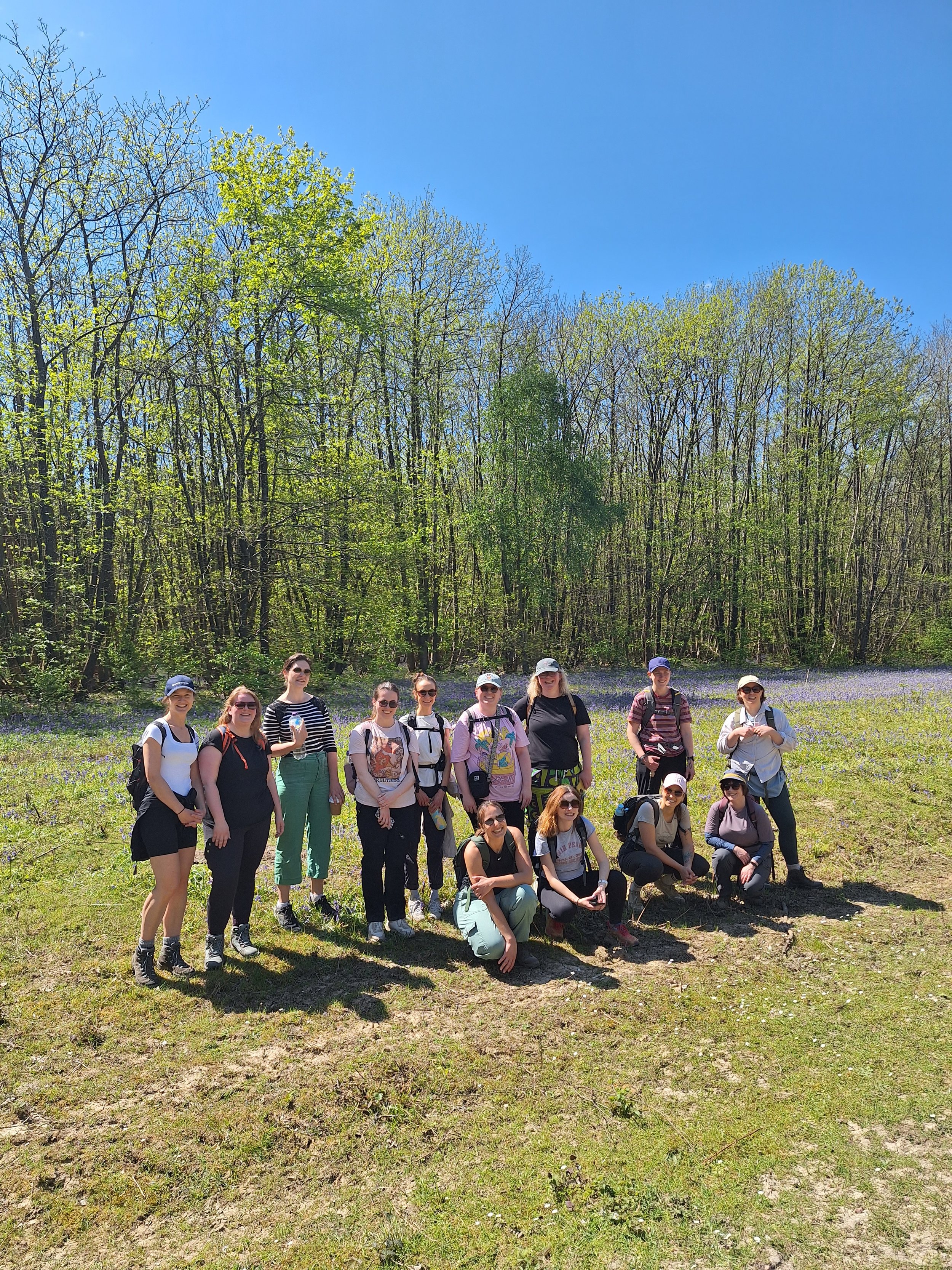 Group of women posing outdoors in a field with purple flowers, with trees and a bright blue sky in the background.