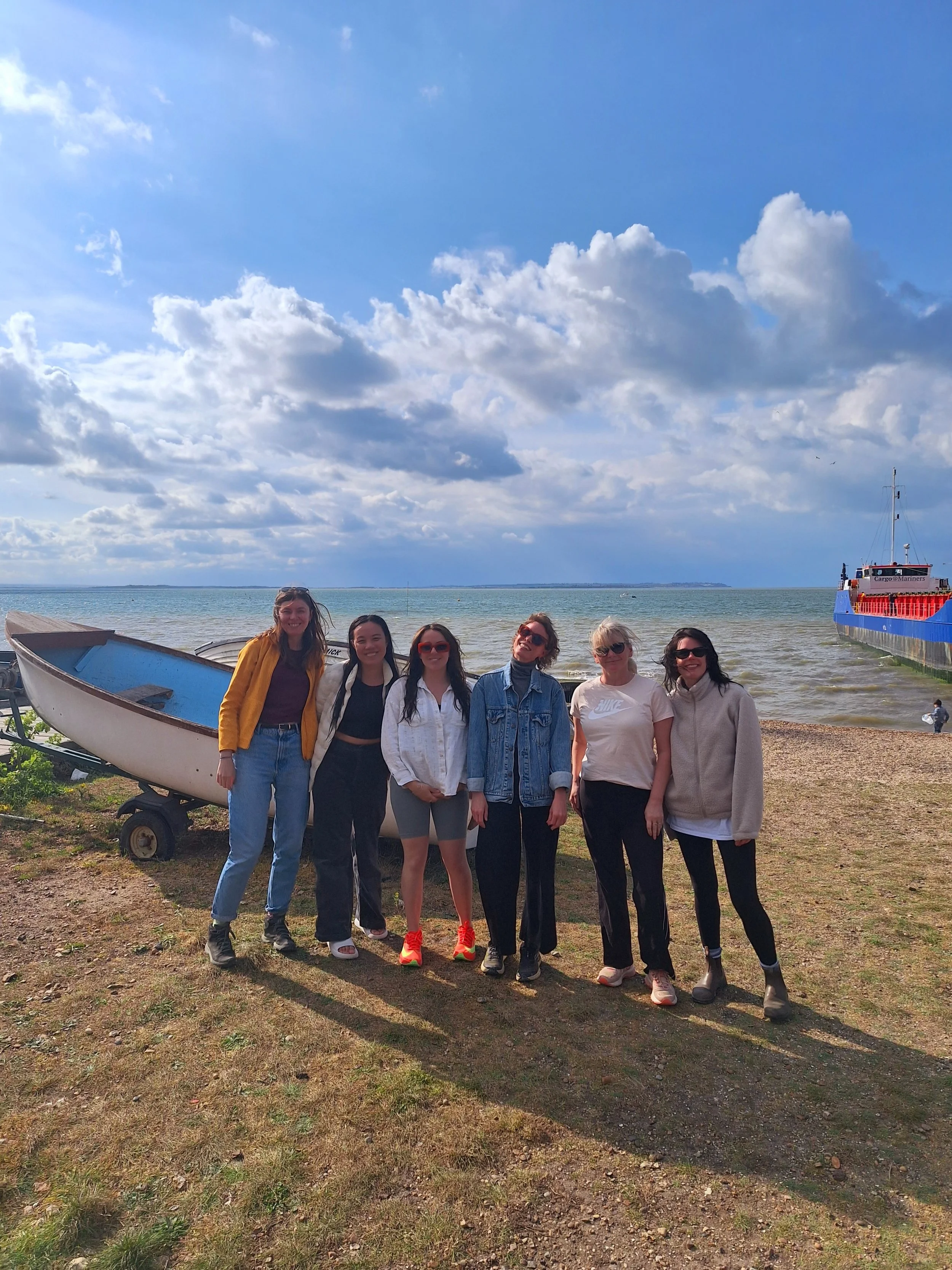 Group of seven women standing on a grassy area near a beach with boats and a blue sky with clouds in the background.