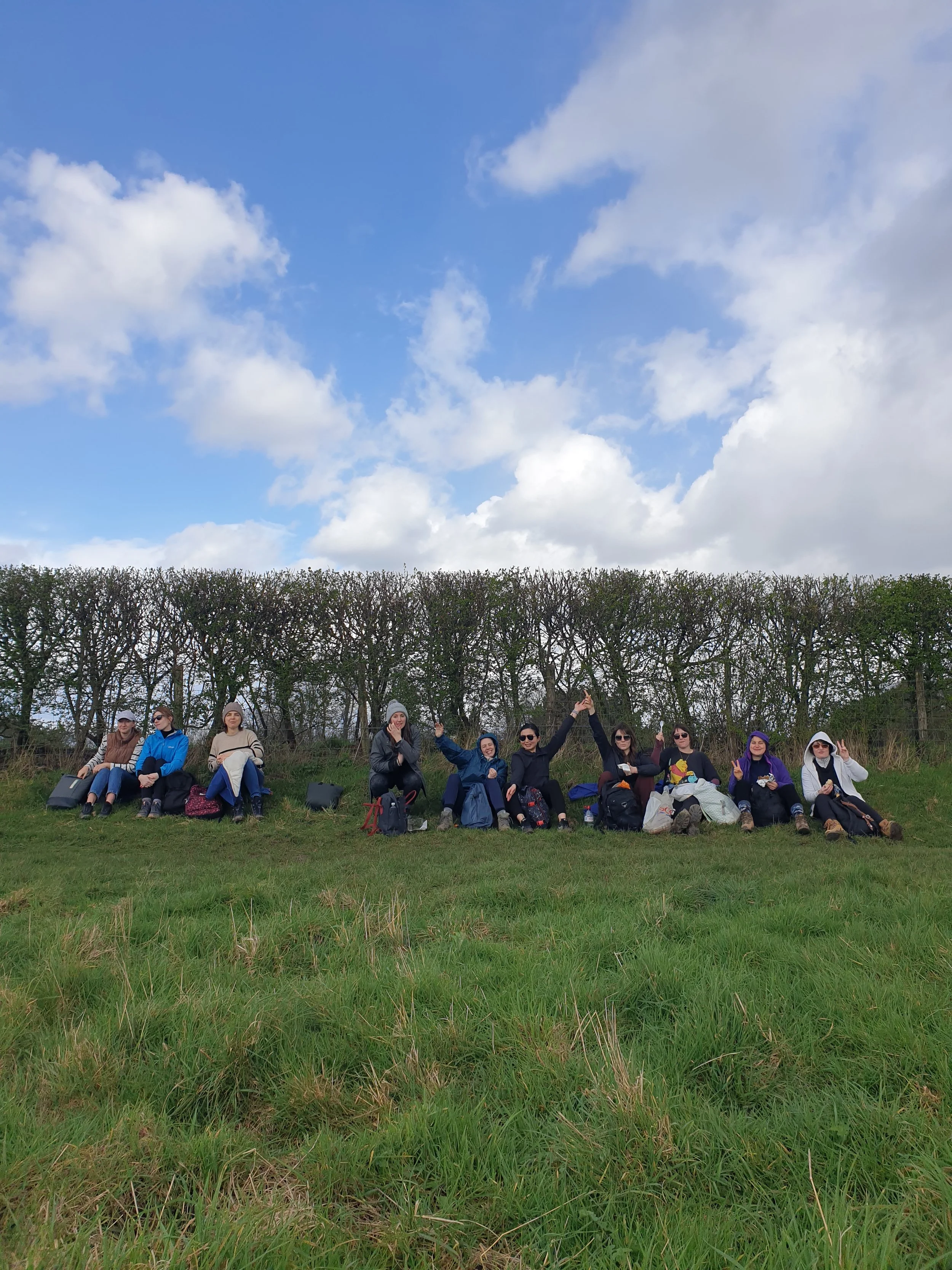 A group of women sitting on the grass outdoors with backpacks and bags, some making peace signs, in front of a row of leafless trees under a partly cloudy sky.