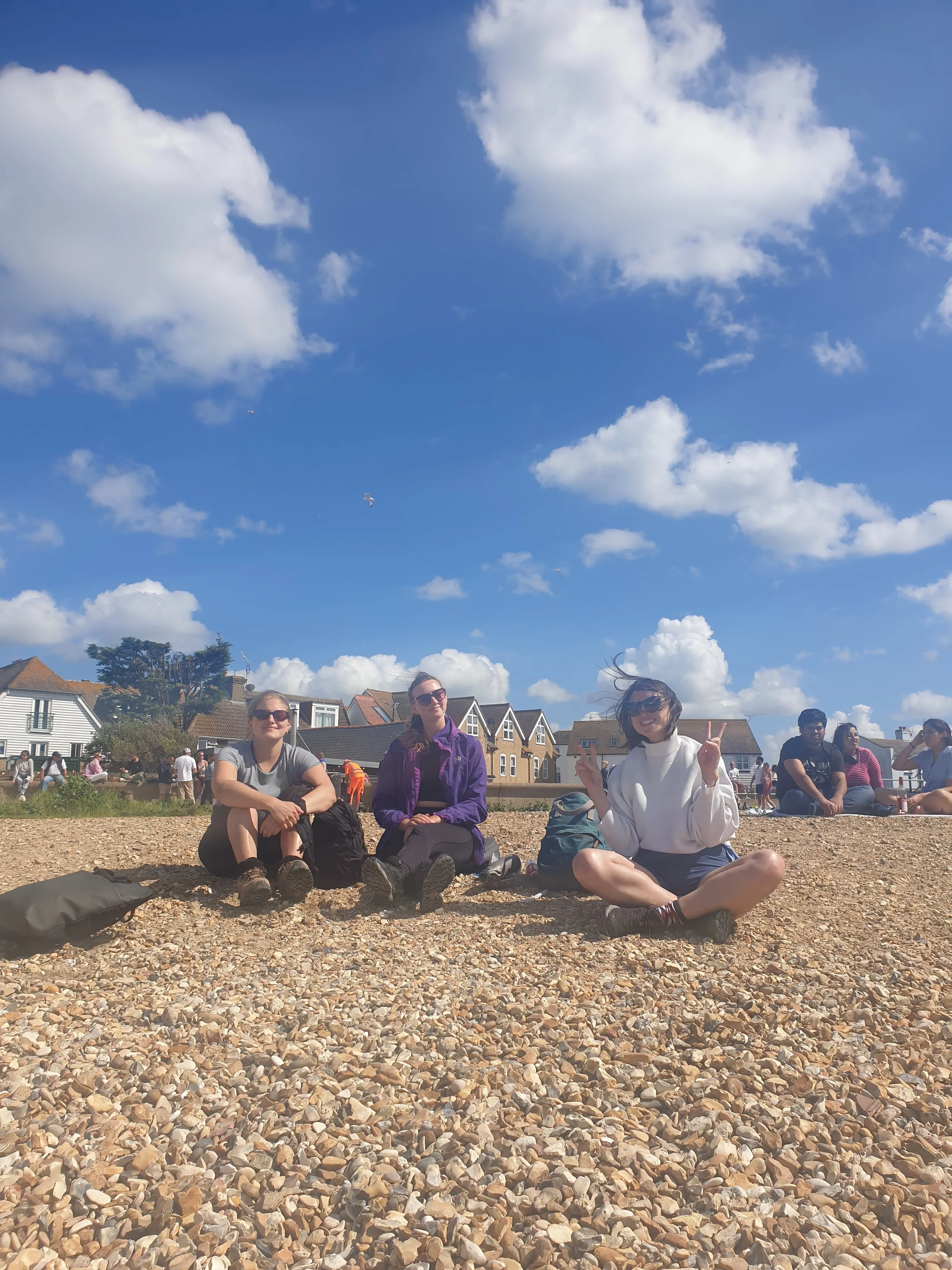 Four women sitting on a pebble beach under a partly cloudy sky, with houses and trees in the background, some people walking around.